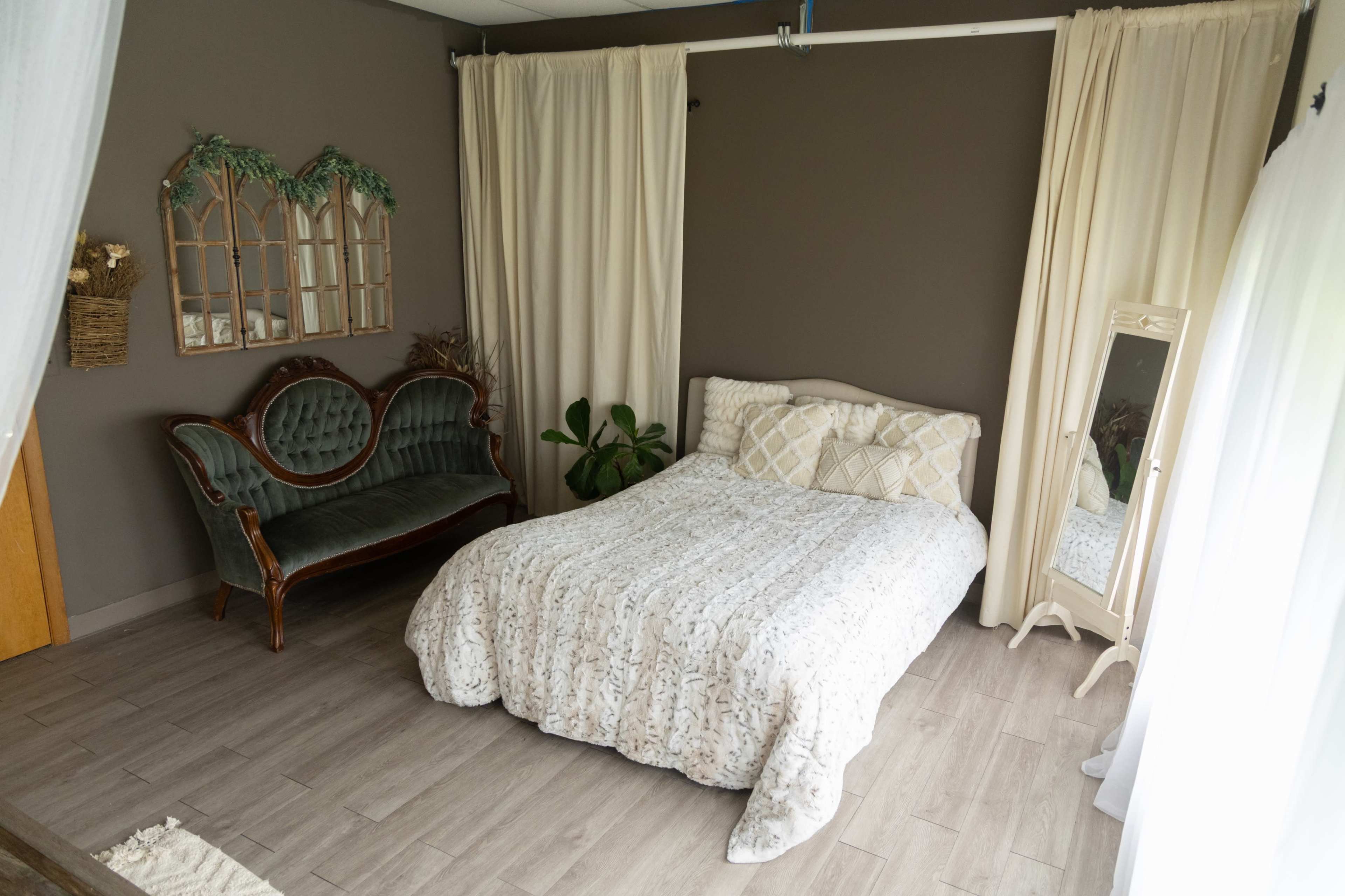 The image shows a bedroom with a bed covered in a patterned white blanket, a vintage sofa, a mirror, and light curtains along the walls.