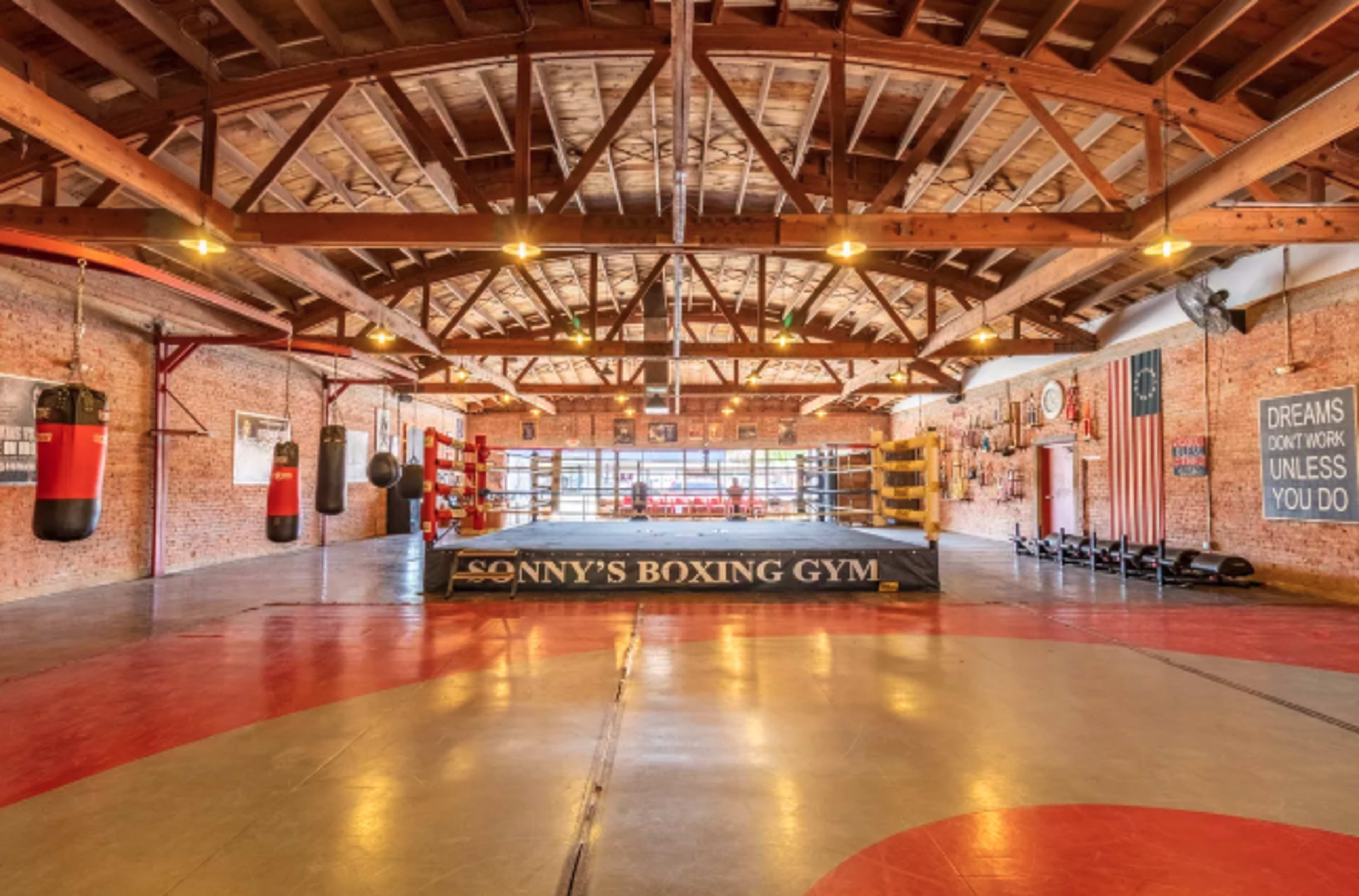The image shows an empty boxing gym with a ring in the center, surrounded by various training equipment and a high wooden ceiling.