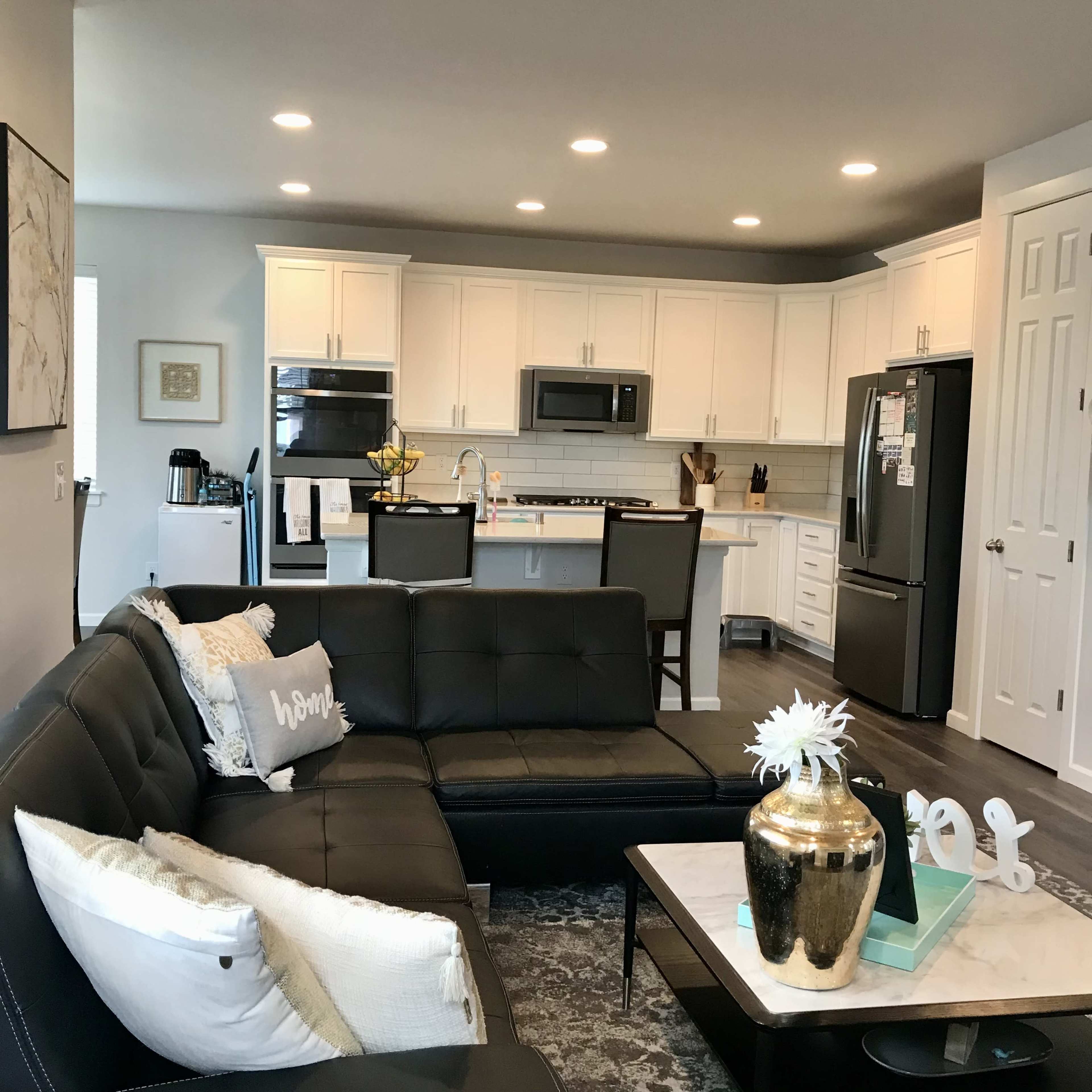 A modern kitchen and living area features a black sectional sofa, white cabinetry, stainless steel appliances, and a central island with seating.