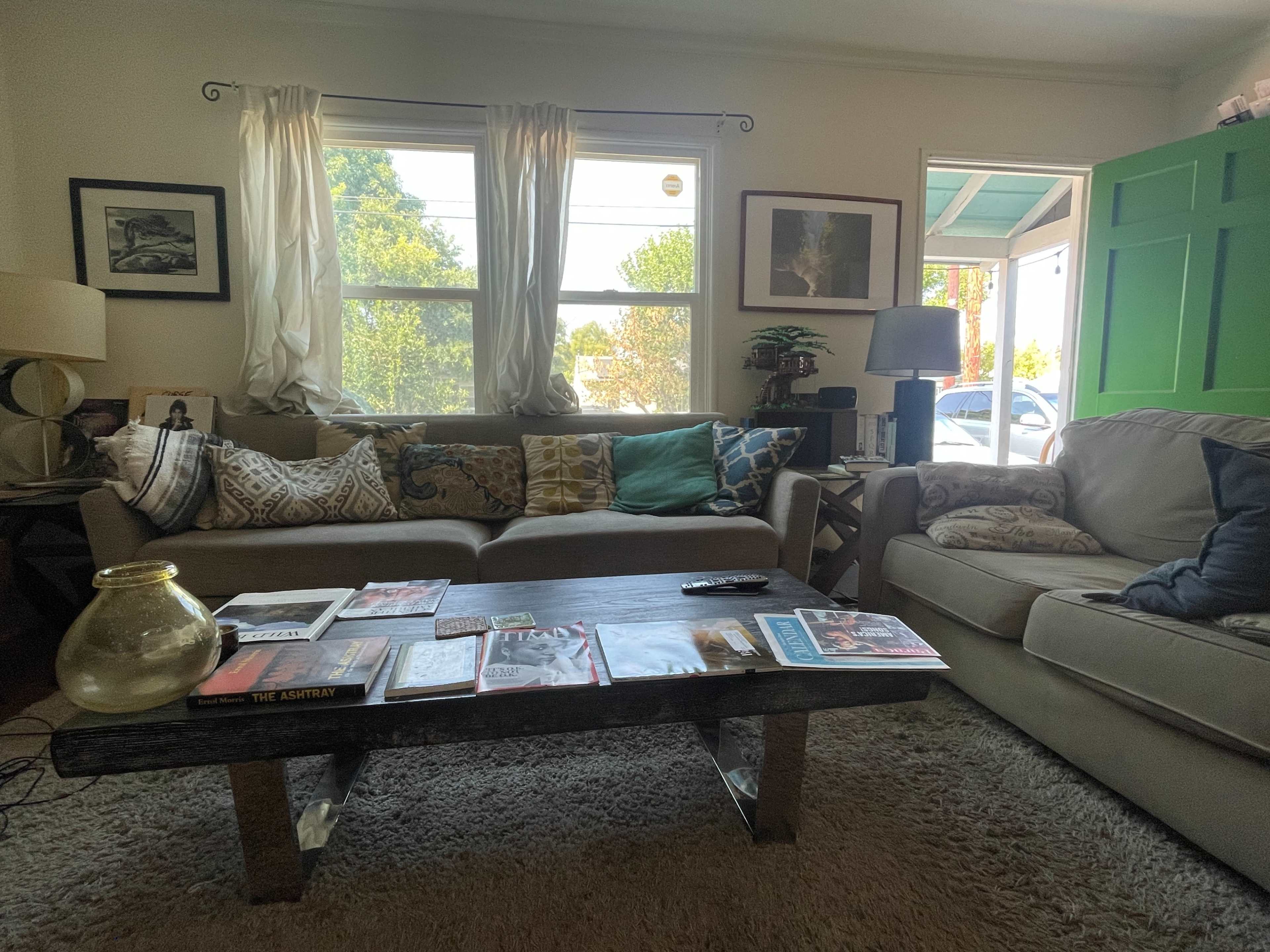 The living room features a comfortable seating area with a coffee table covered in magazines and a large window allowing natural light to fill the space.