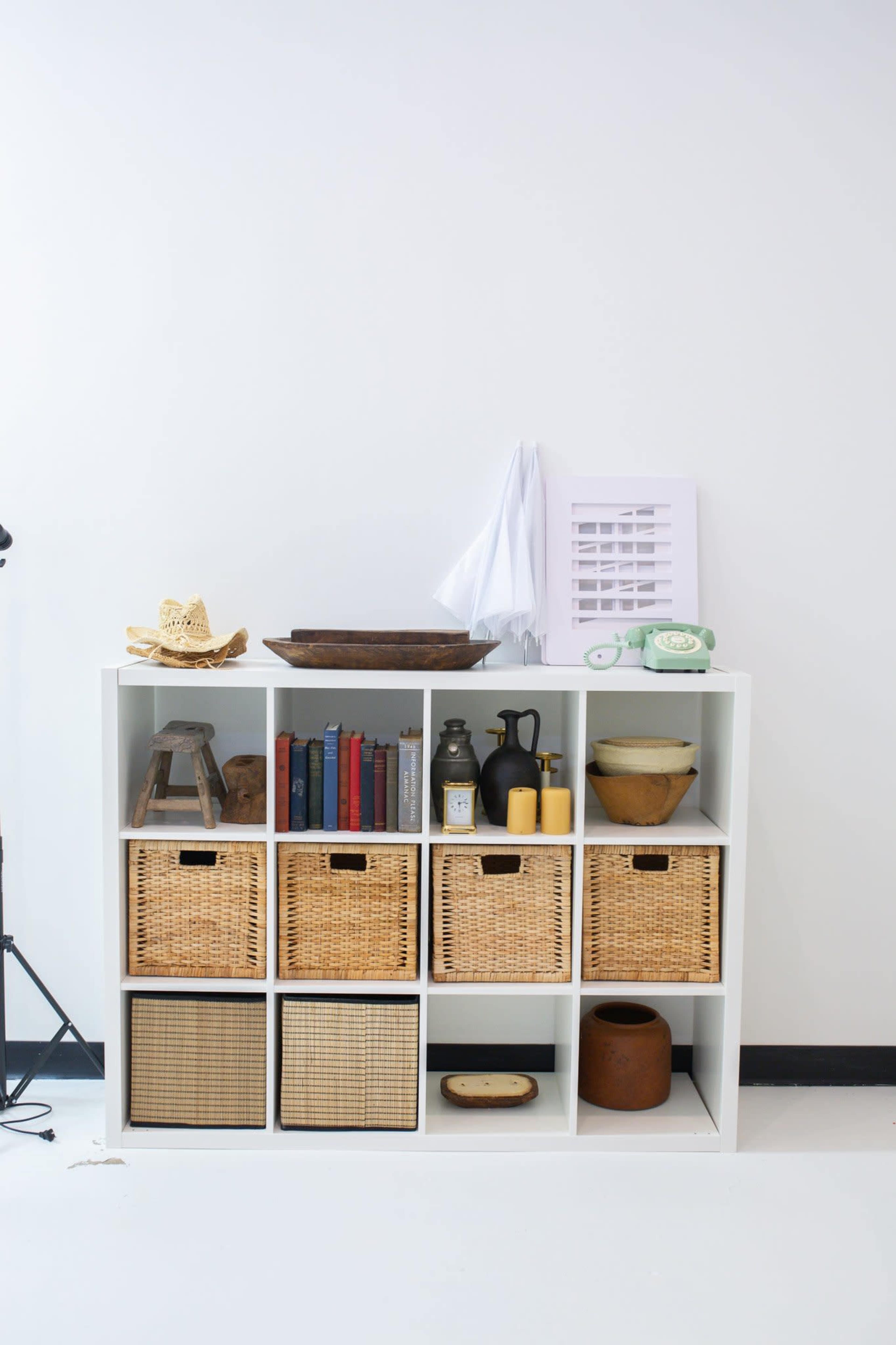 A white bookshelf displays various items, including books, woven baskets, kitchenware, and decorative objects against a plain wall.