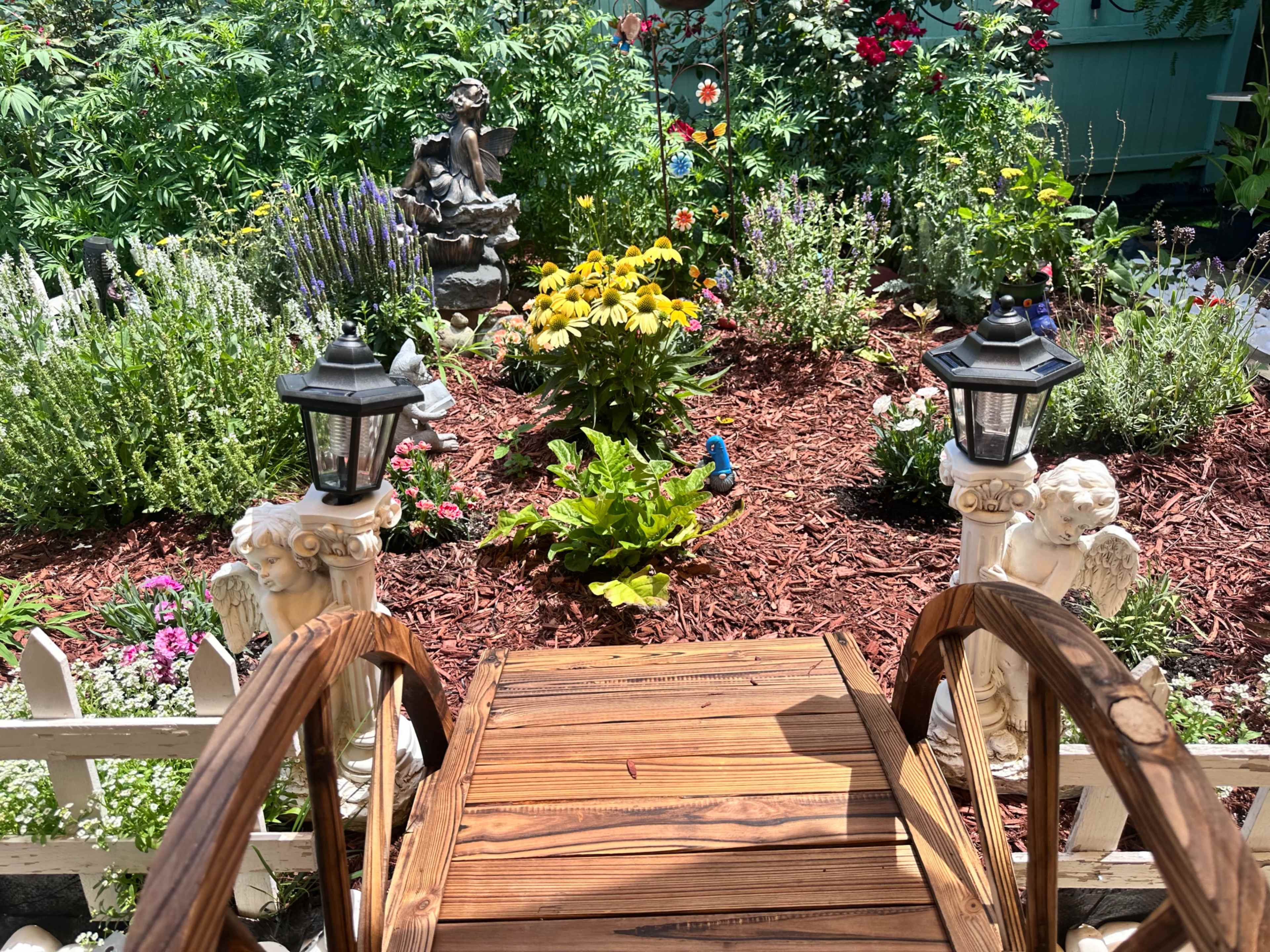 The image shows a well-maintained garden featuring a variety of colorful flowers, a statue, and decorative lampposts, with a wooden bridge in the foreground.