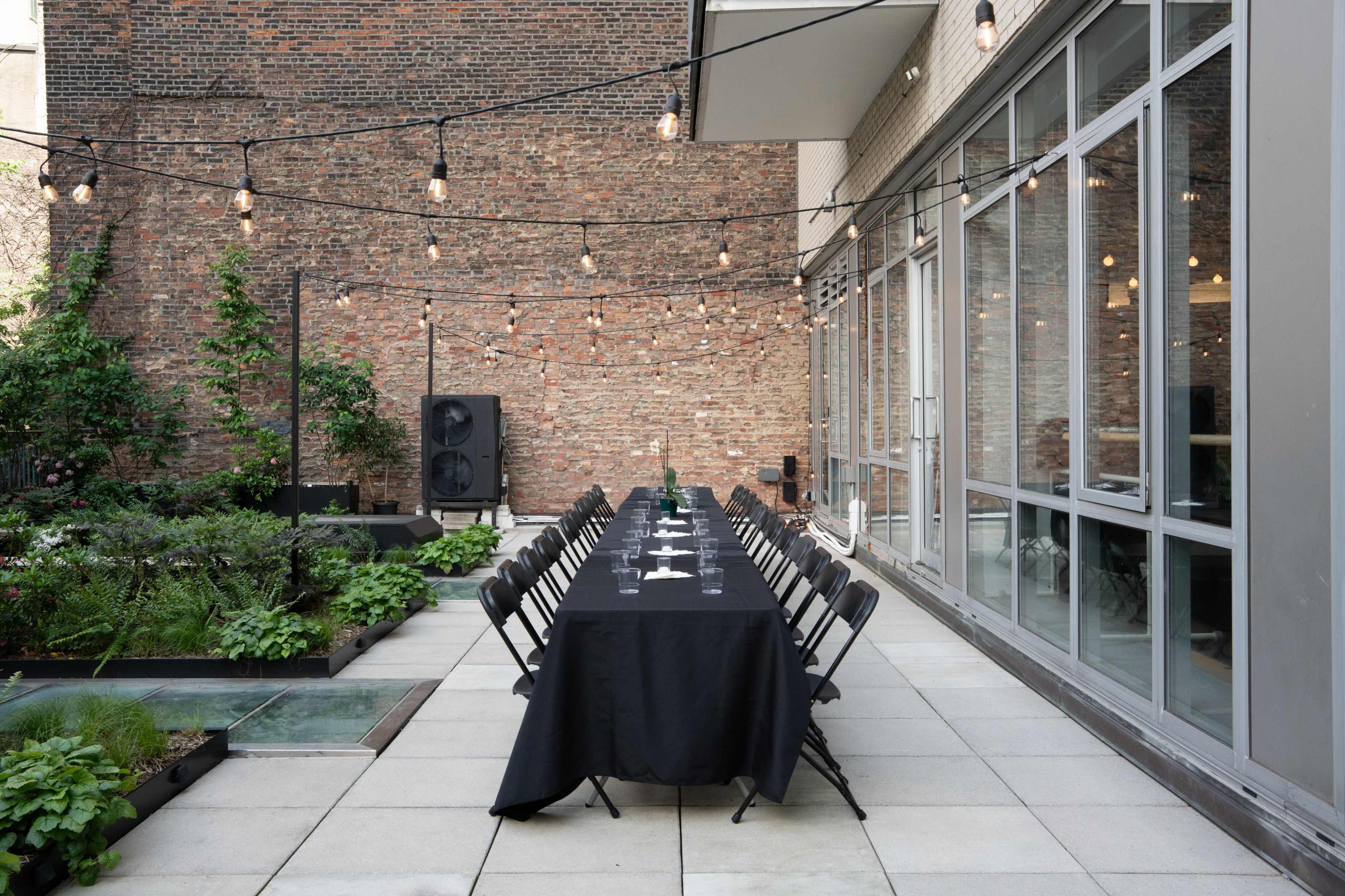 A long table with black tablecloth and chairs is set up in an outdoor space surrounded by greenery and a brick wall, with string lights overhead.