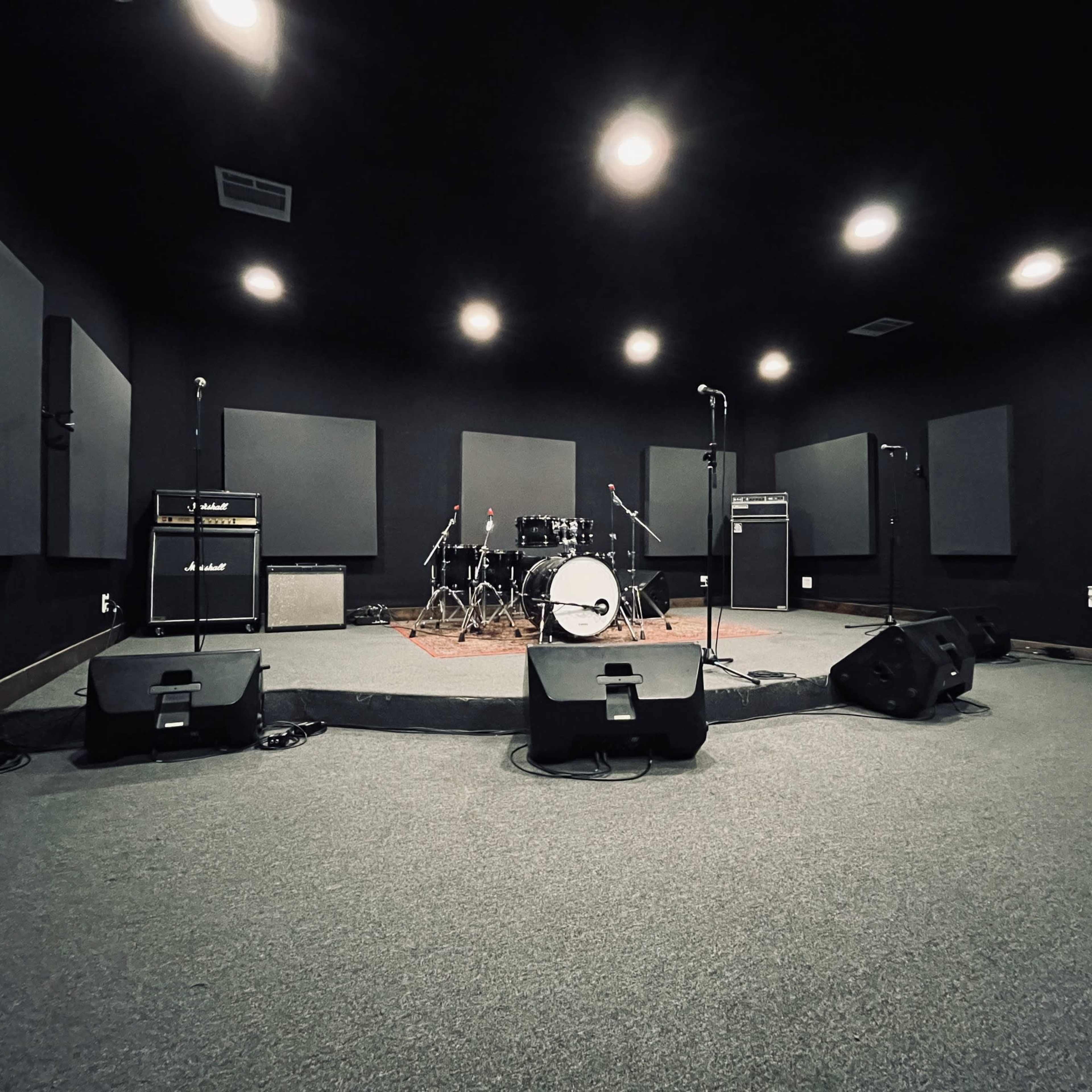 A soundproof rehearsal room features a drum kit, amplifiers, and microphone stands, with black walls and ceiling lights.