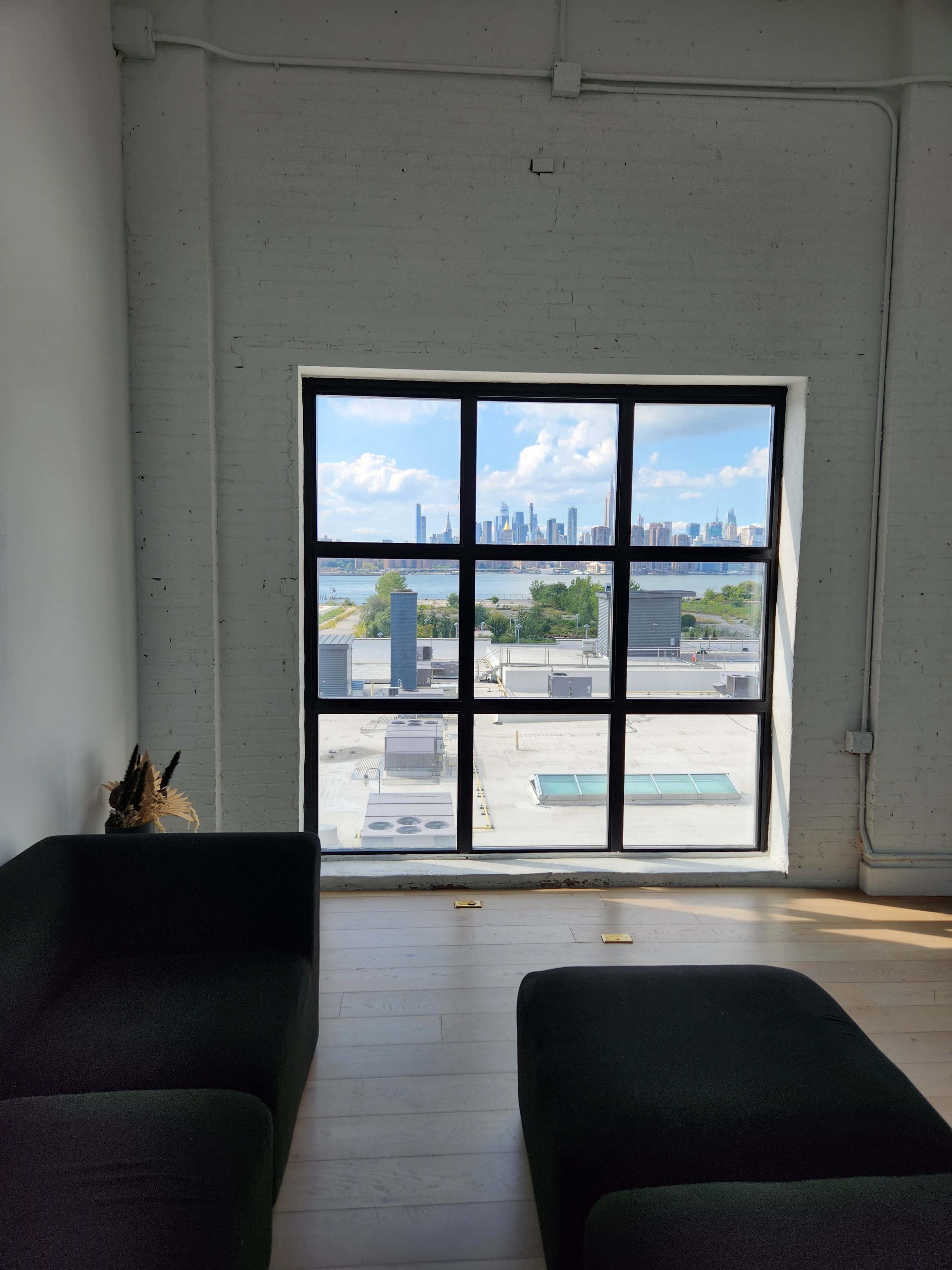 A large window frames a view of a city skyline against a clear sky, with a modern interior space featuring dark furniture in the foreground.