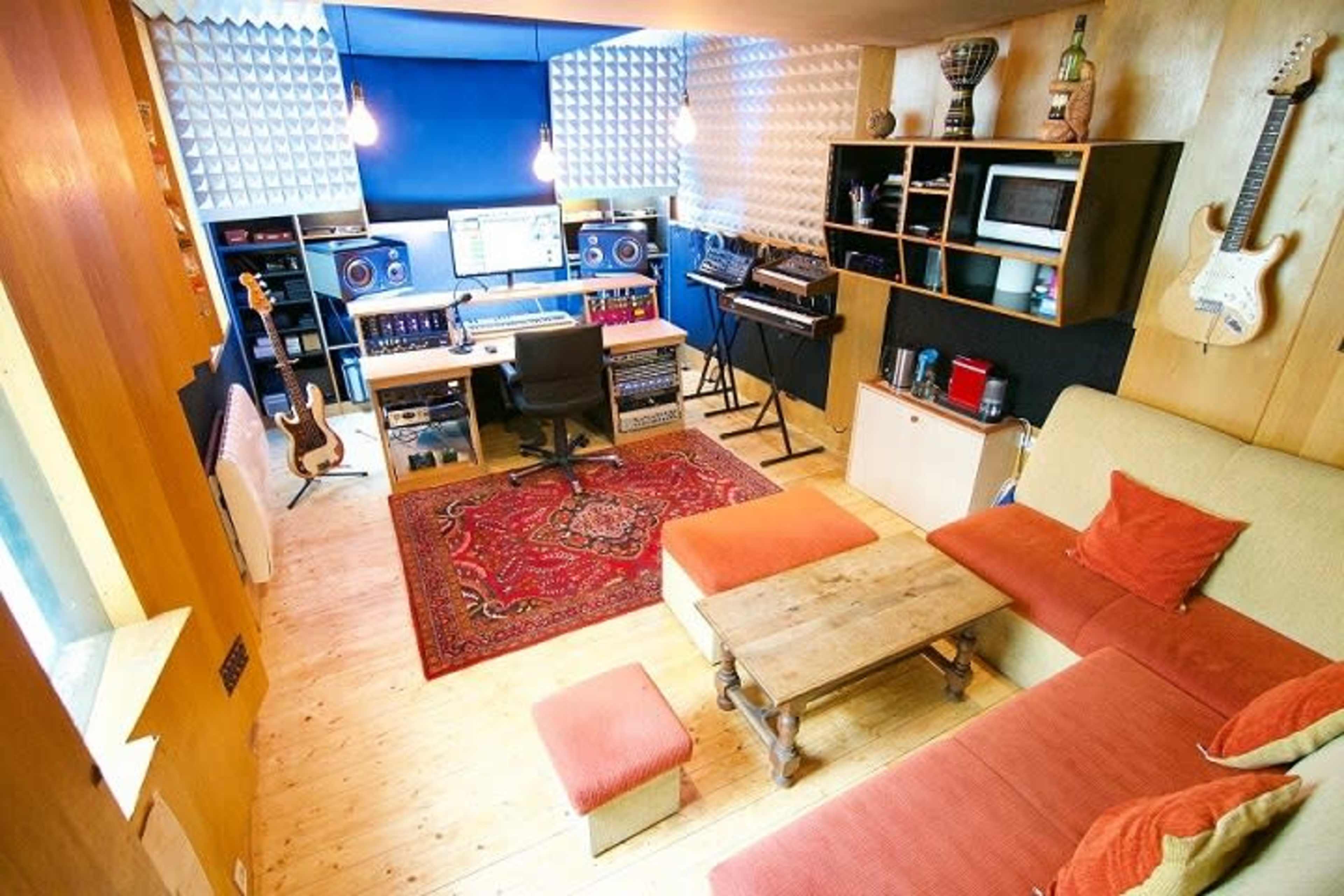 The image shows a home recording studio with a mix of instruments, sound equipment, and a cozy seating area, all featuring wooden accents and soundproofing panels.