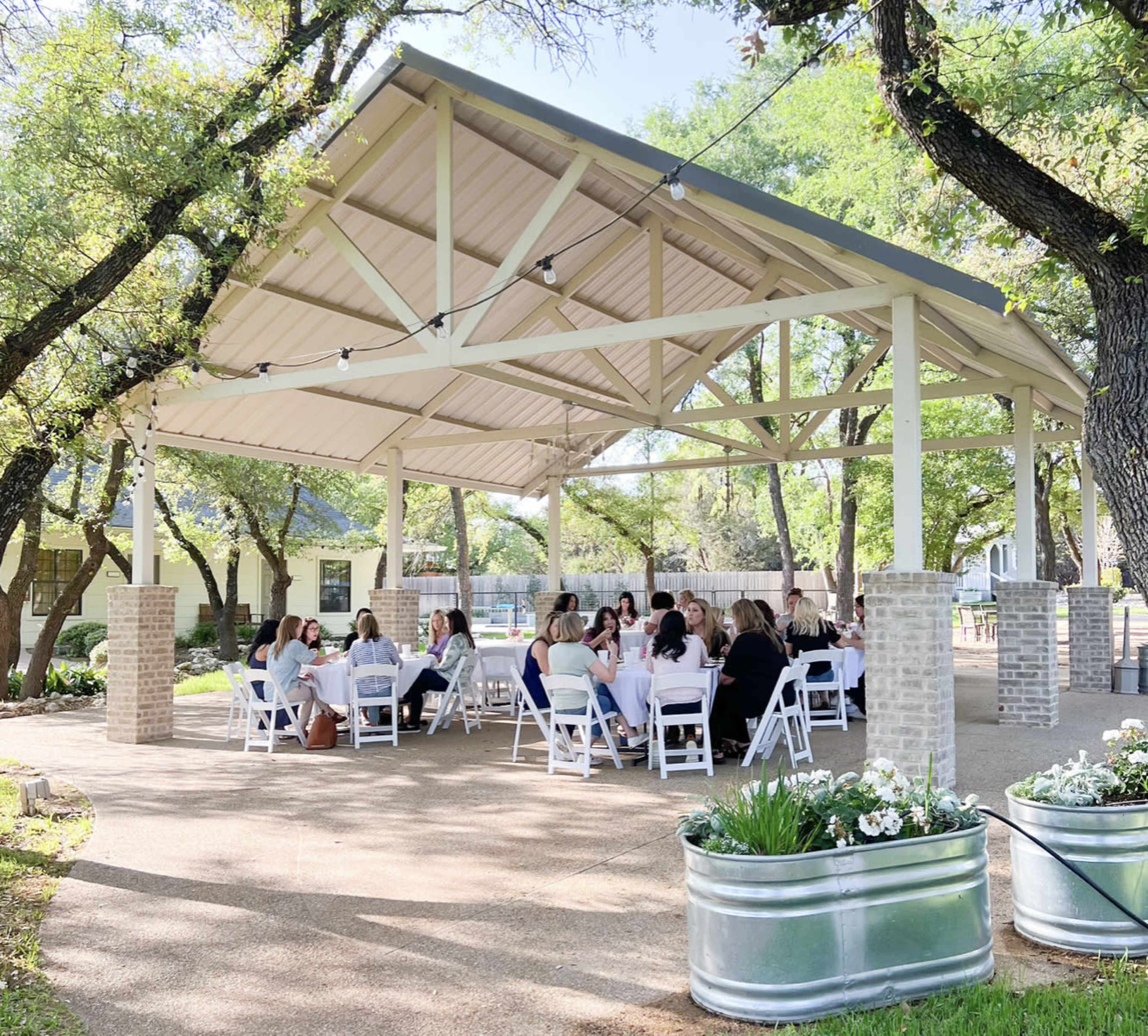 A group of people sits around a circular arrangement of tables beneath a covered outdoor pavilion surrounded by trees and greenery.
