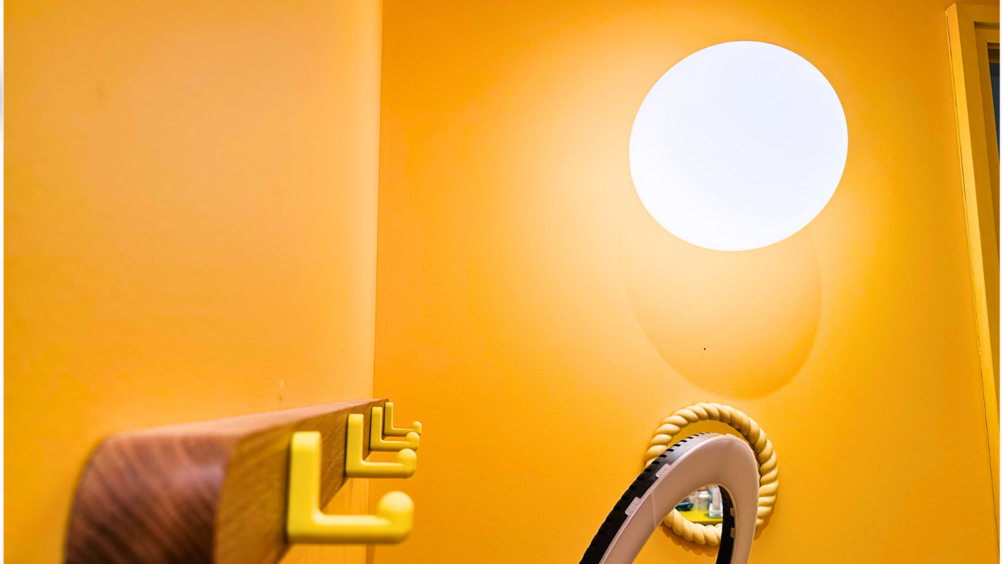 The image shows a brightly colored yellow wall with a round light fixture and a wooden coat rack with several hooks.