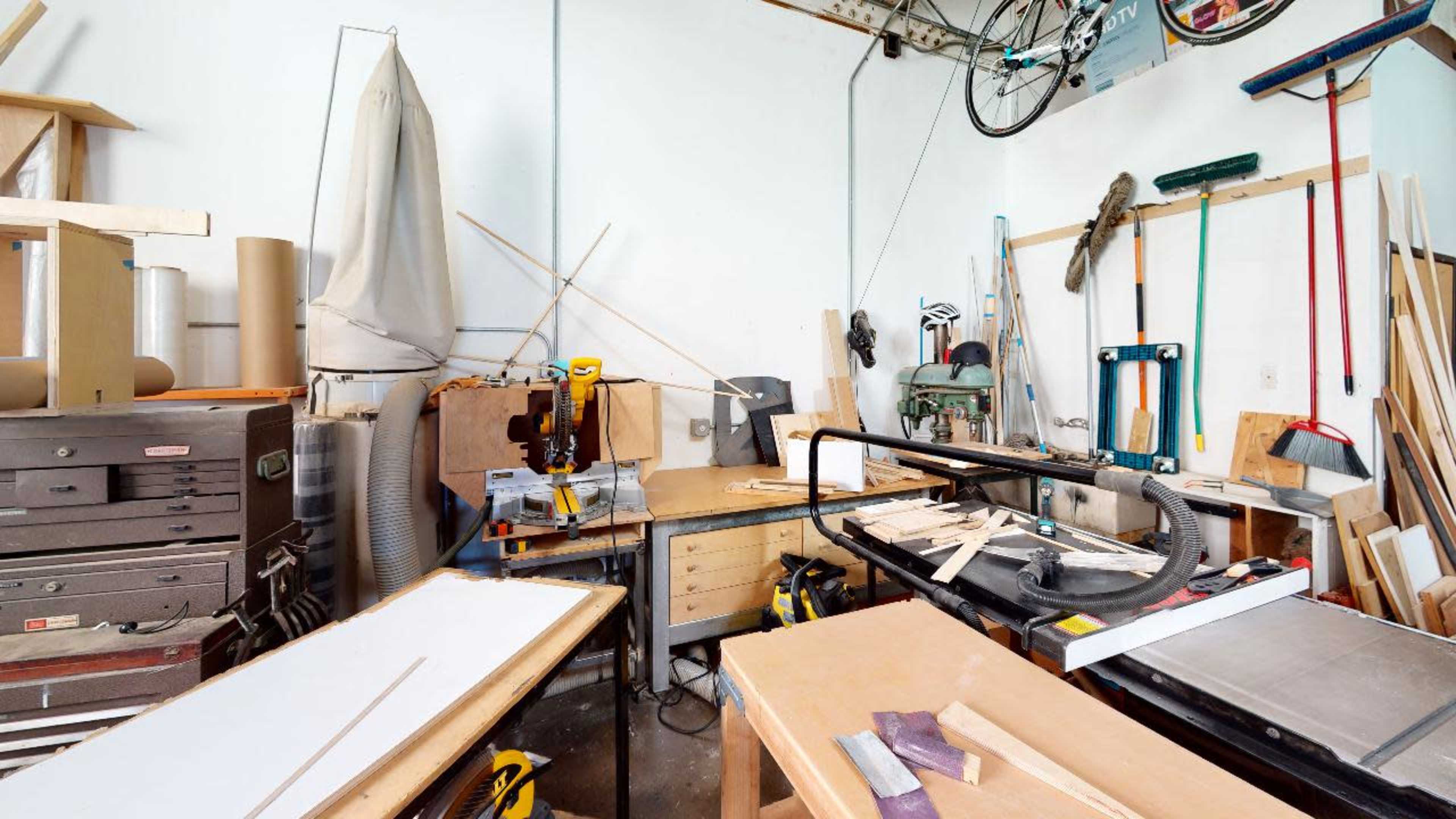A cluttered workshop with various woodworking tools, a workbench, and piles of lumber.