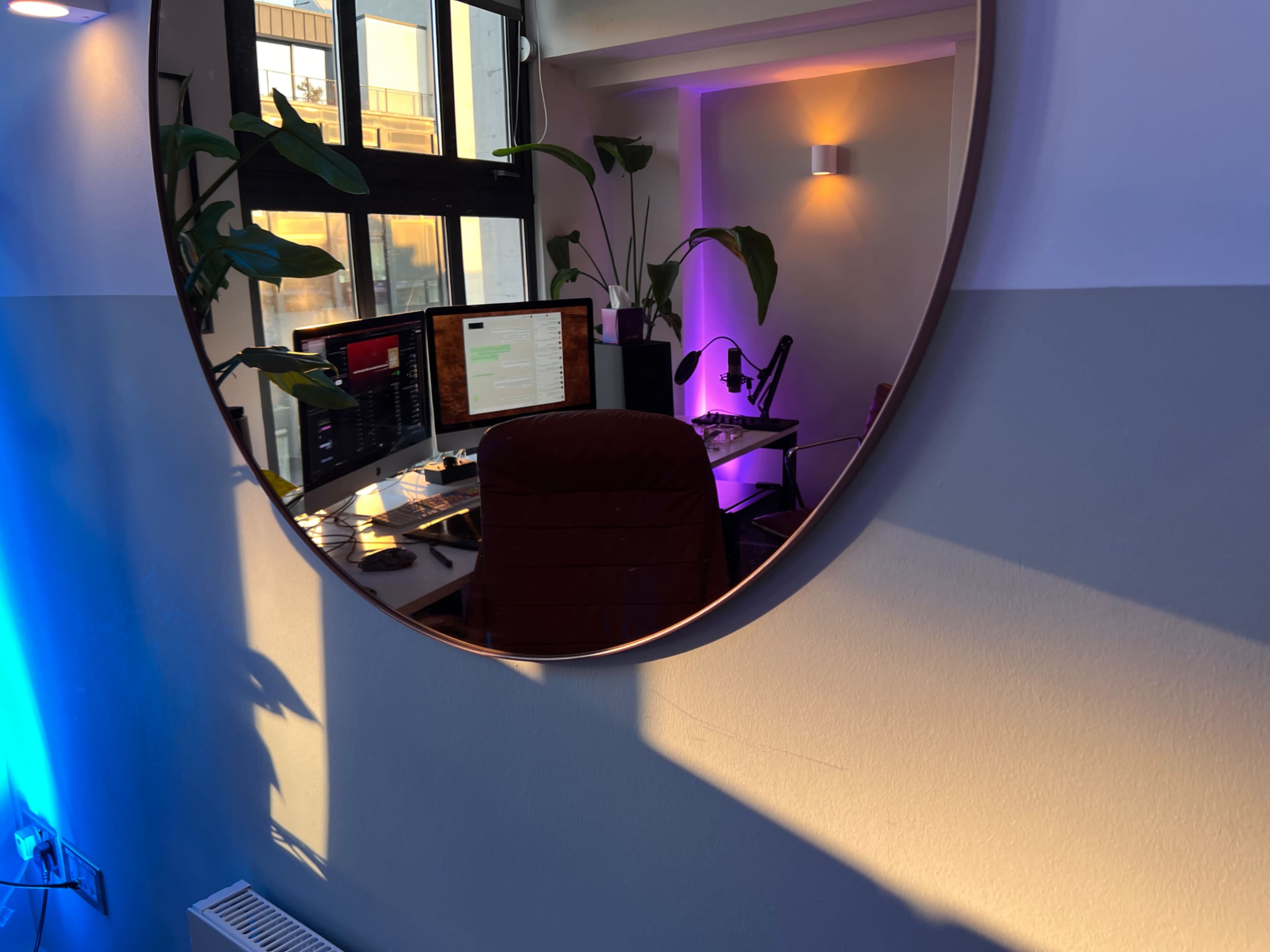 The image shows a large round mirror reflecting a workspace with computer monitors, a microphone, and indoor plants, illuminated by colored lights.