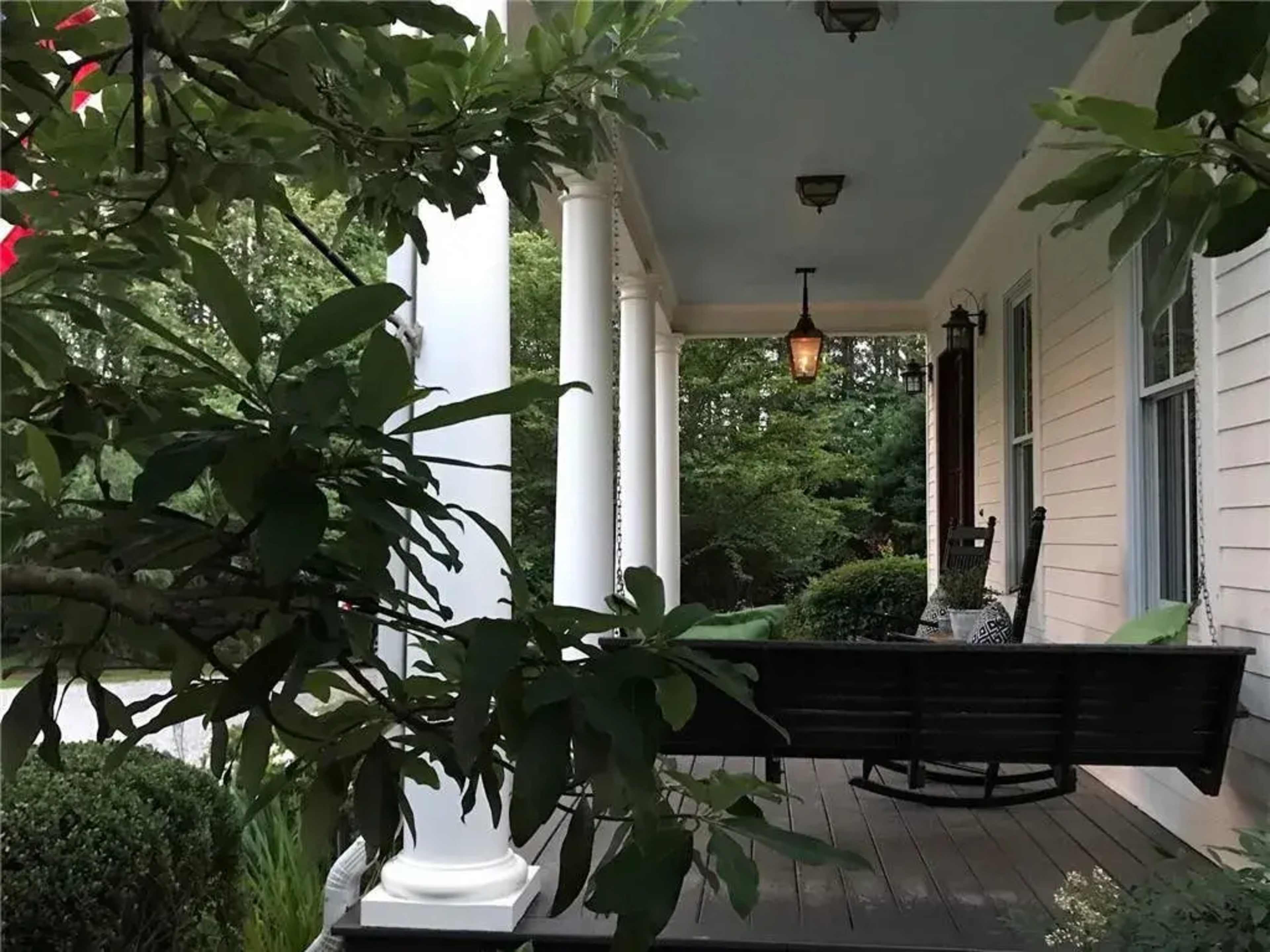 A porch features a swing attached to a column with a hanging lantern above, surrounded by greenery.