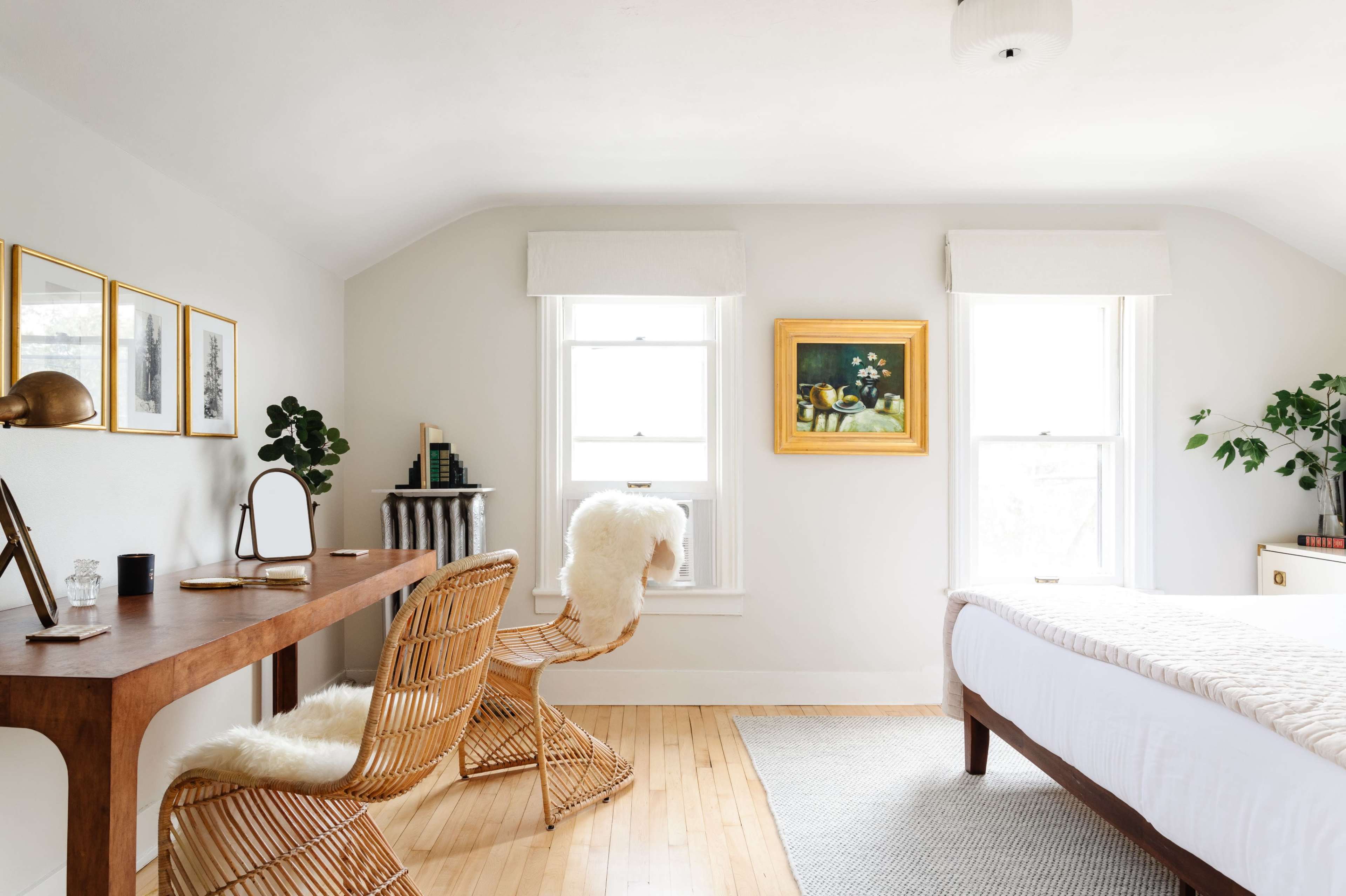 A well-lit bedroom features a wooden desk with a chair, a bed with a light quilt, and artwork displayed on the walls.