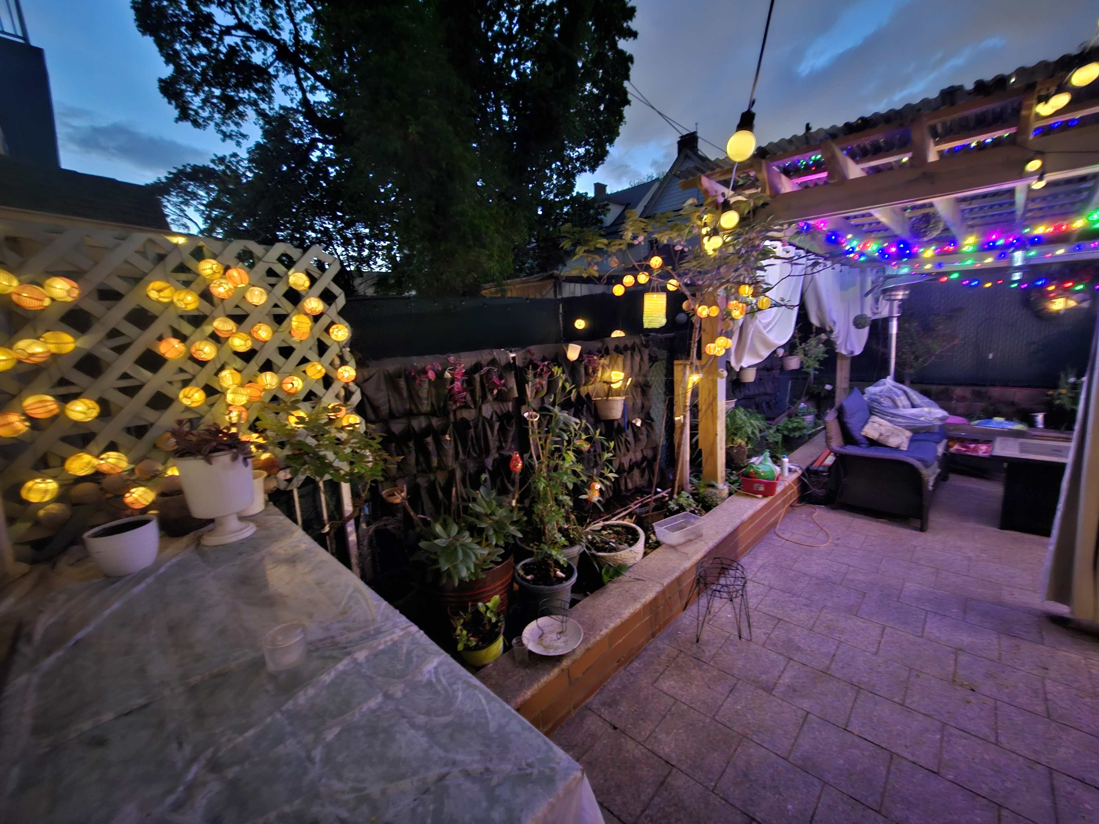 A patio scene at dusk features string lights illuminating potted plants and a seating area under a pergola.