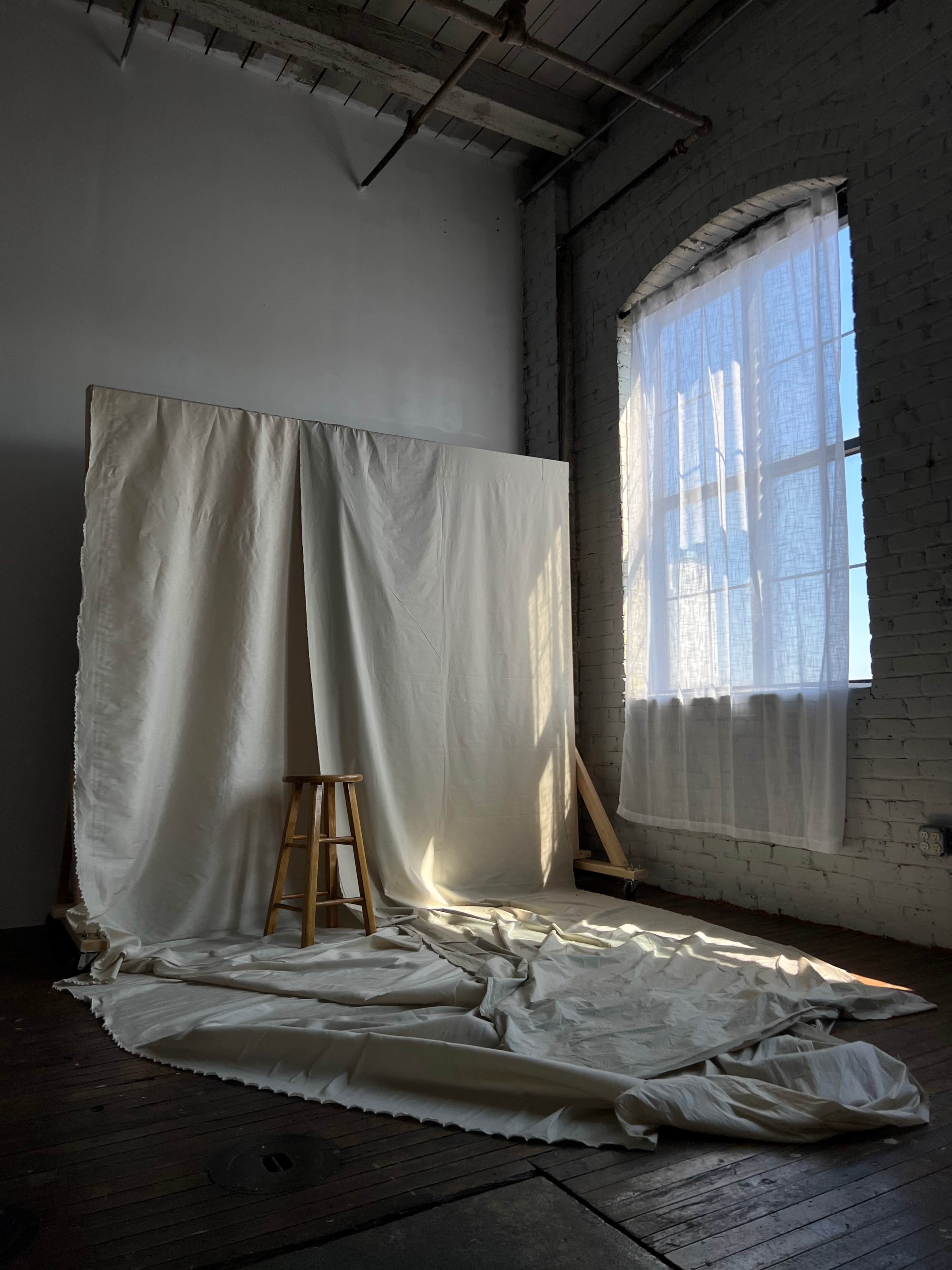 A simple room features a large white fabric backdrop and a wooden stool beside a window with sunlight streaming through.