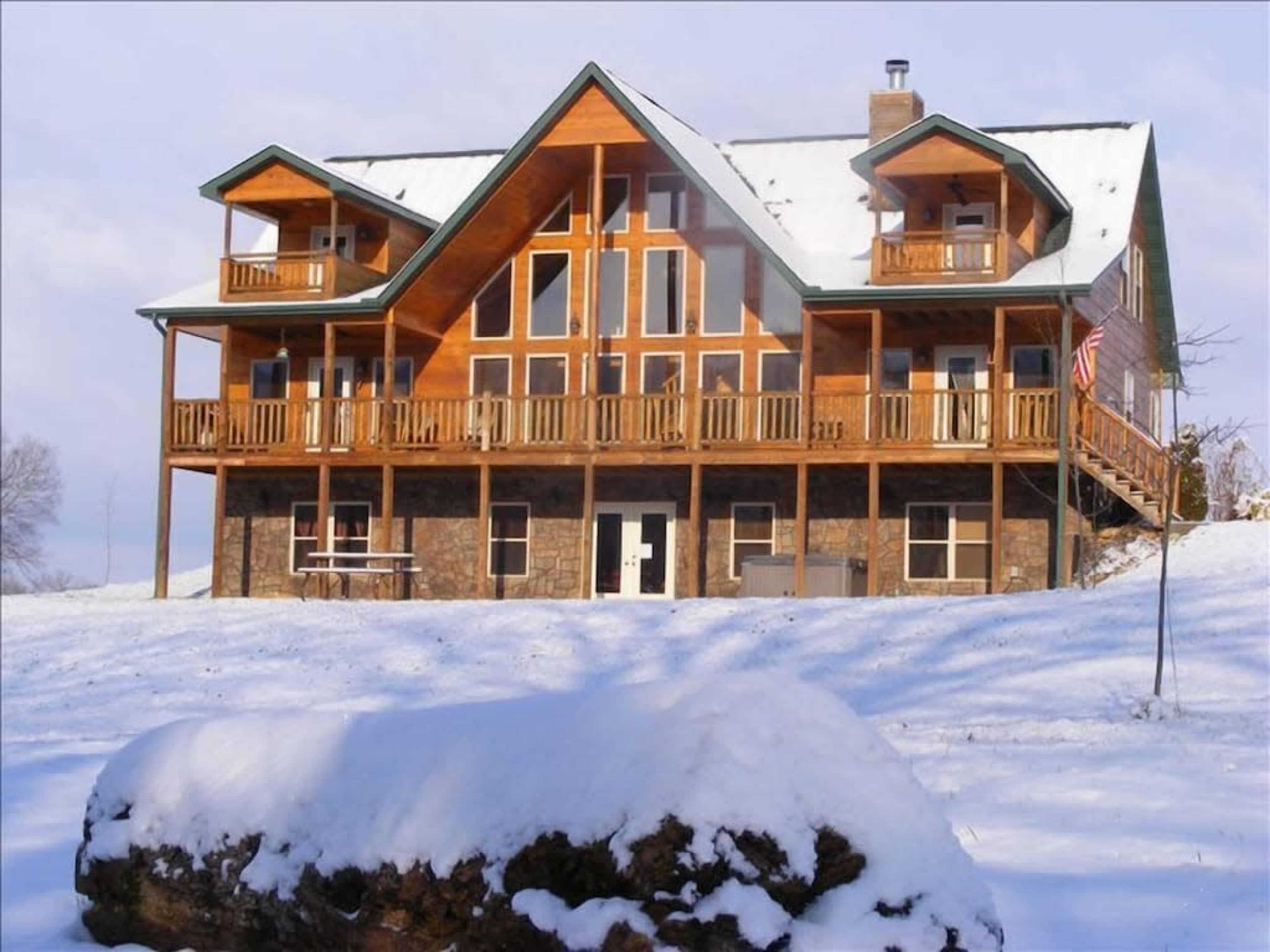 A large multi-level wooden house with stone accents sits in a snowy landscape, featuring multiple balconies and large windows.