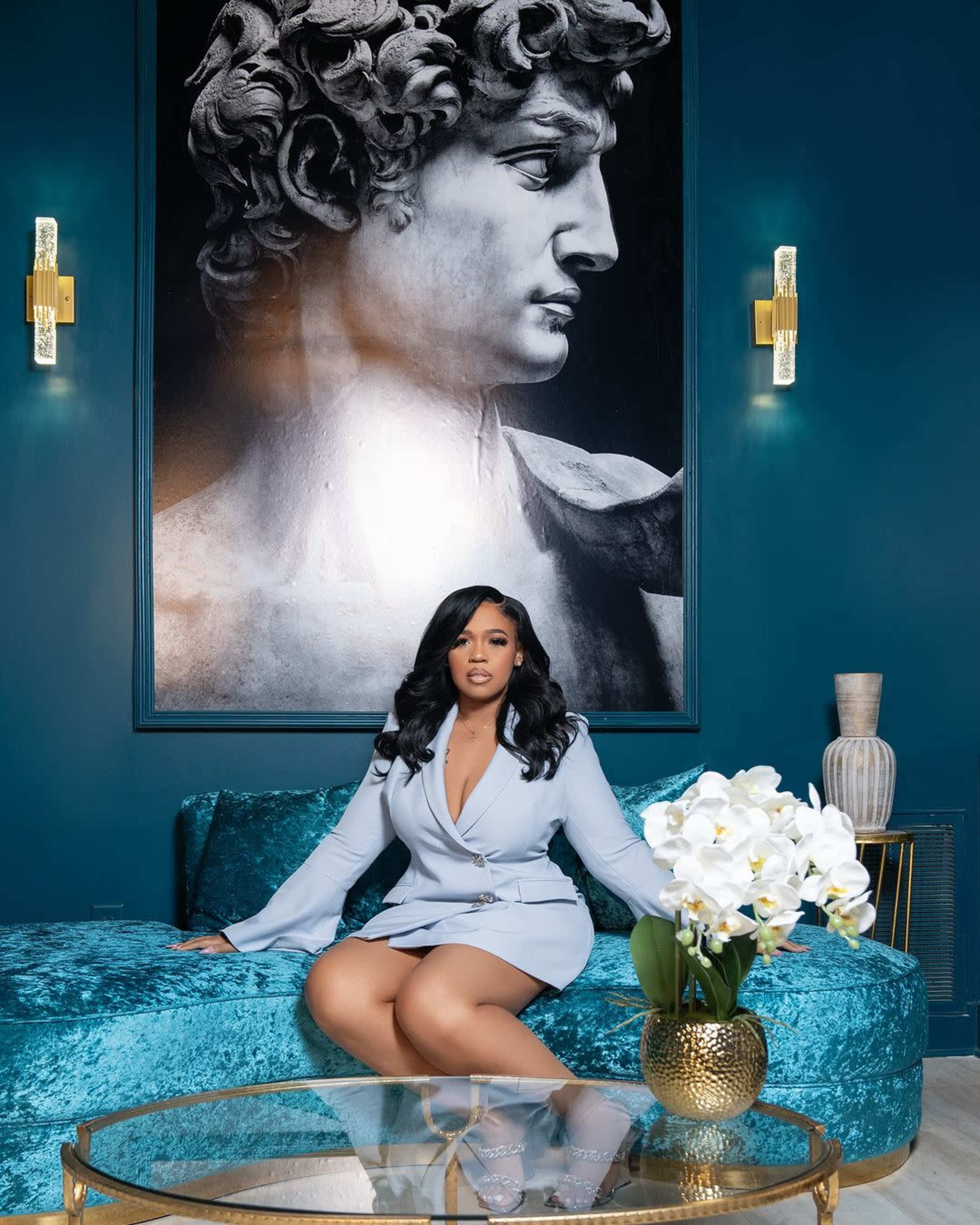 A woman in a light blue outfit sits on a teal couch in front of a large black-and-white portrait of a classical sculpture.
