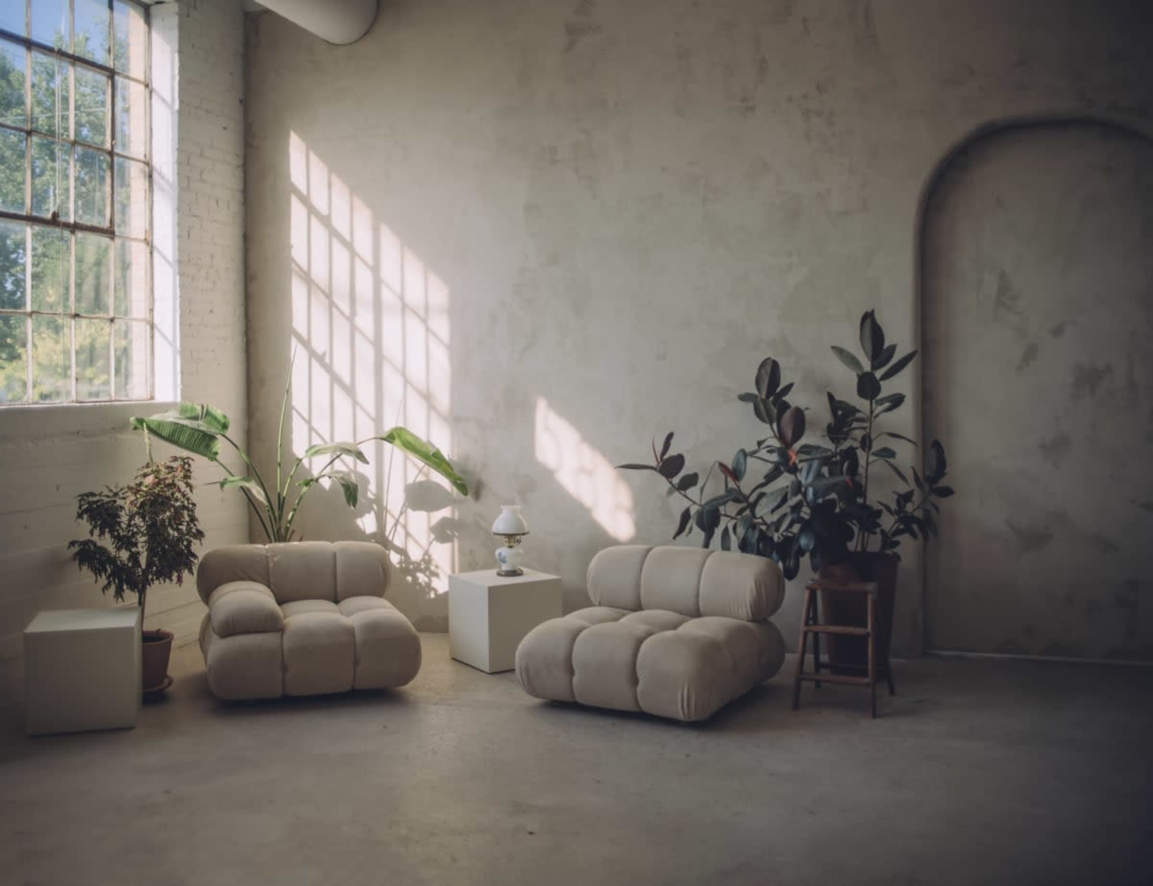 A minimalist interior space with two beige couches, a small white table, a lamp, and potted plants against a textured wall and large windows.