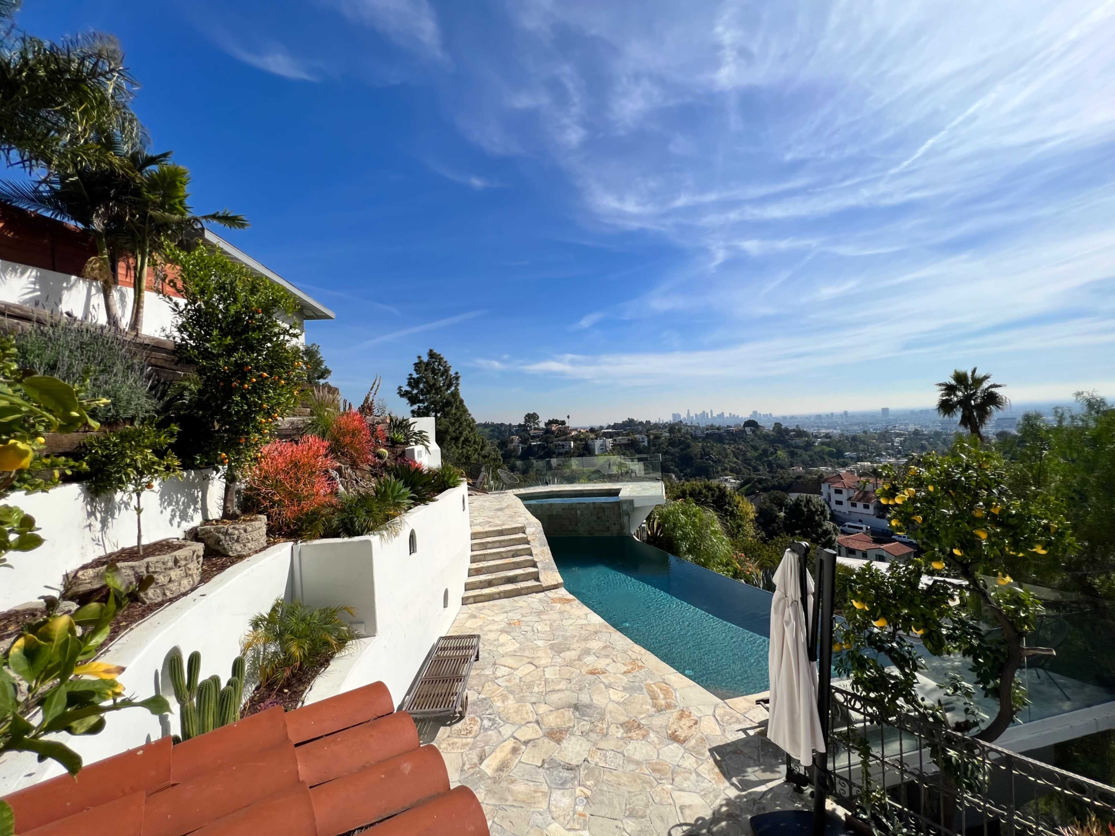 The image shows a hillside pool area with stone pathways, lush greenery, and city views under a clear blue sky.
