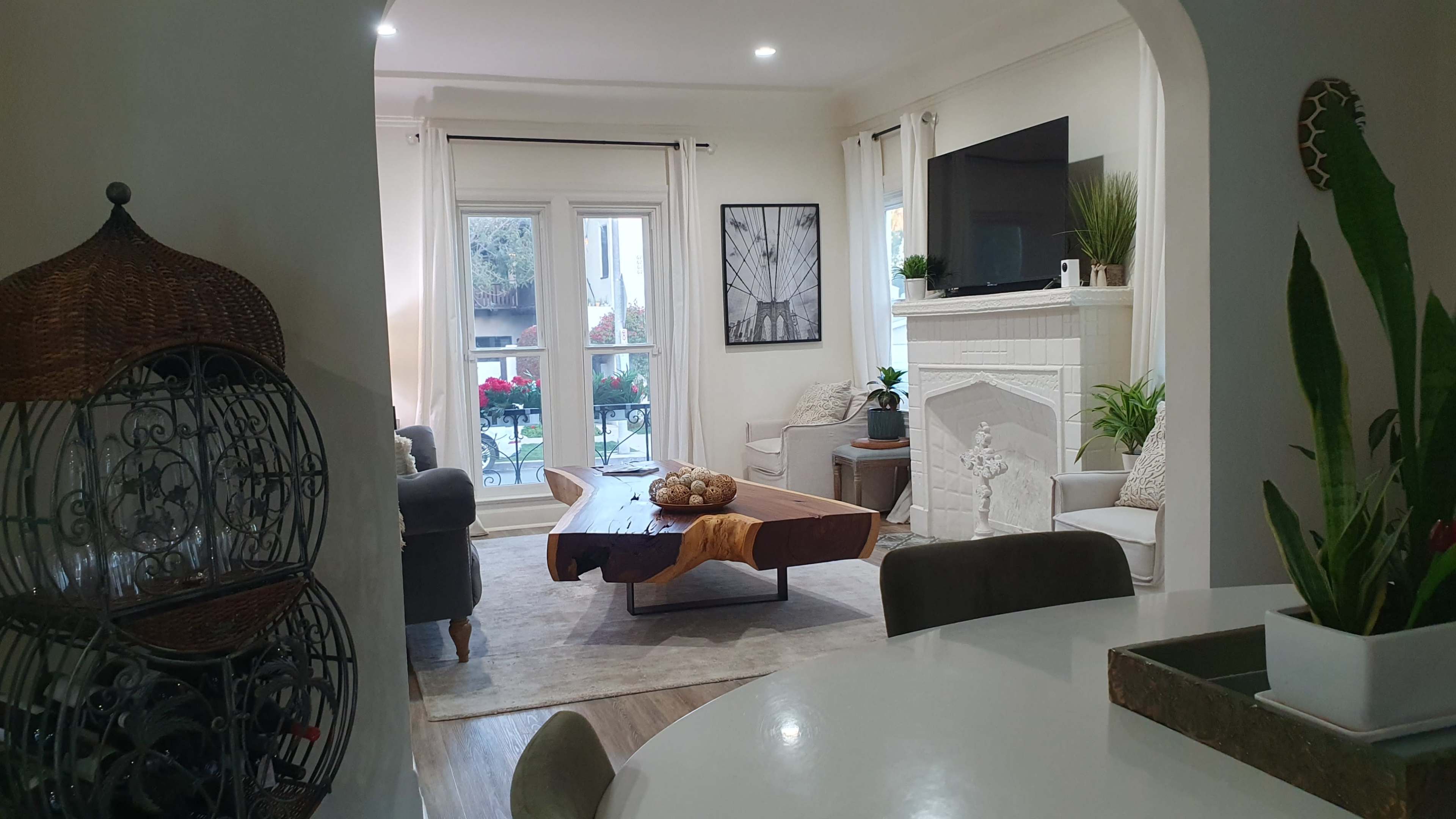 The image shows a modern living room with a wooden coffee table, a white fireplace, and large windows overlooking a garden.