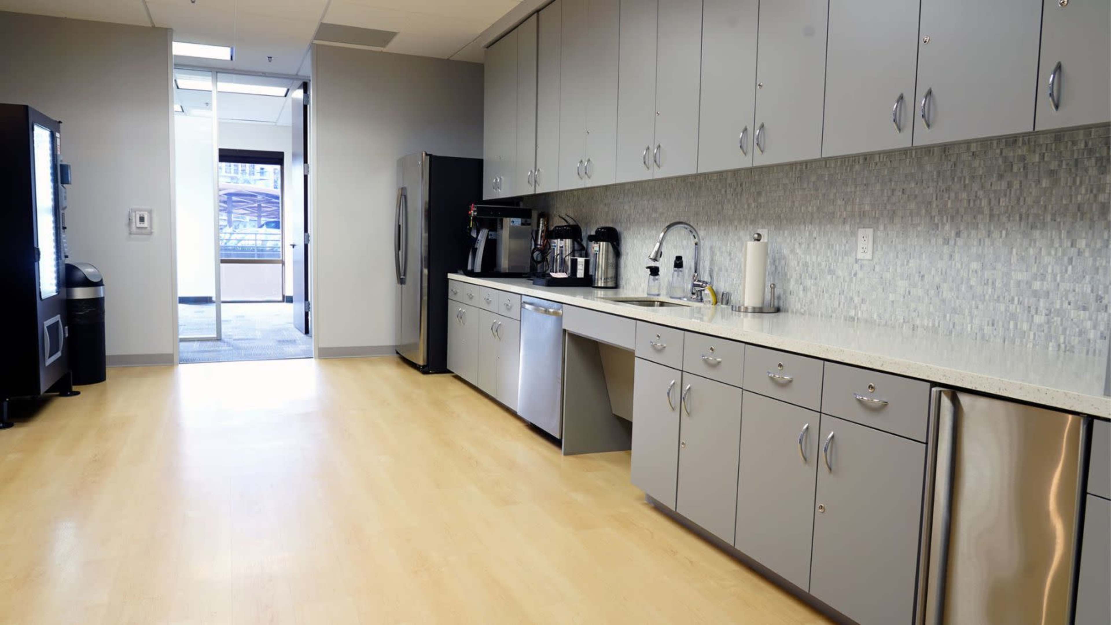 The image shows a modern kitchen area with gray cabinetry, a coffee machine, and appliances, featuring a large counter and an open door leading to an outdoor space.