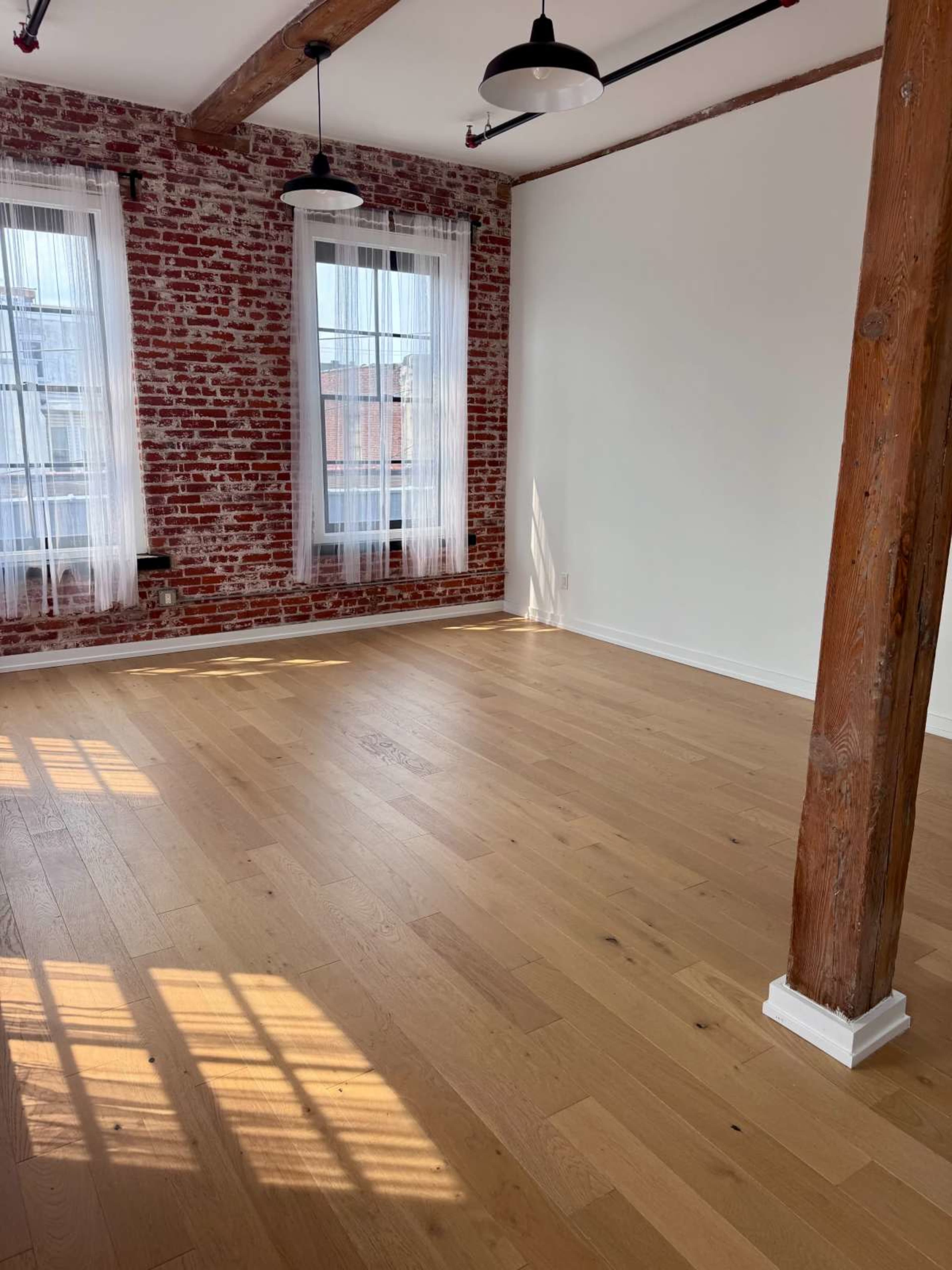 The image shows an empty room with exposed brick walls, large windows with sheer curtains, and wooden flooring.
