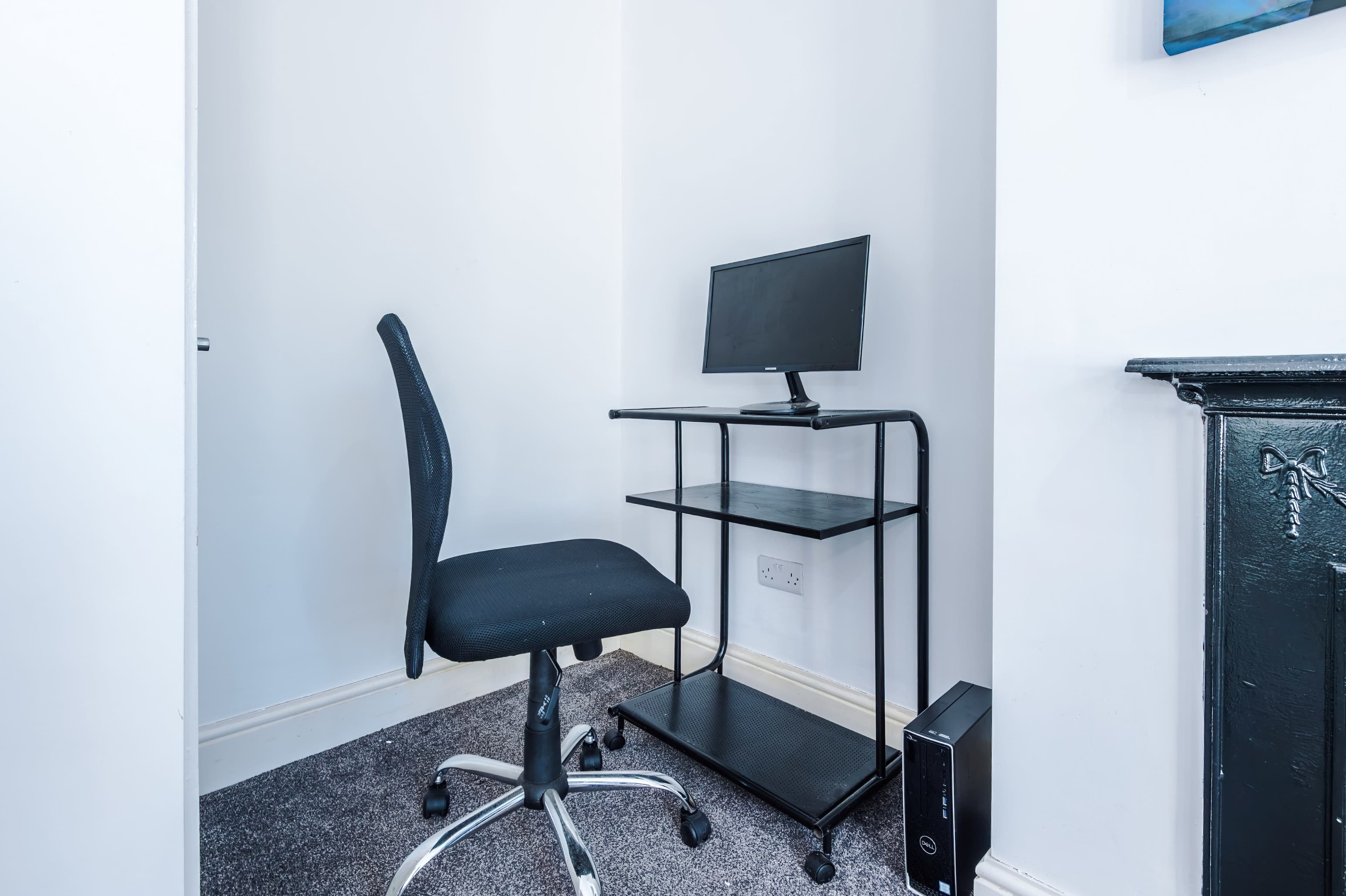 A small office space features a black rolling chair, a computer monitor on a metal stand, and a desktop computer tower against a white wall.