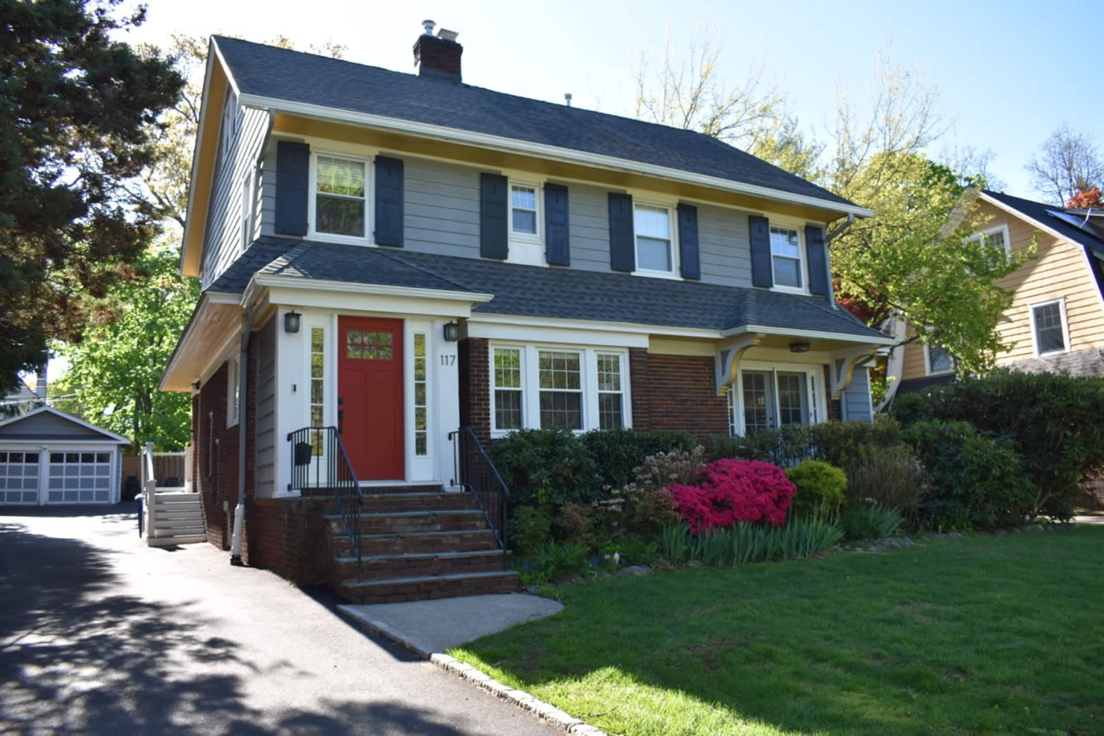The image shows a two-story house with a brick and grey siding exterior, featuring a red front door and surrounded by greenery and blooming flowers.
