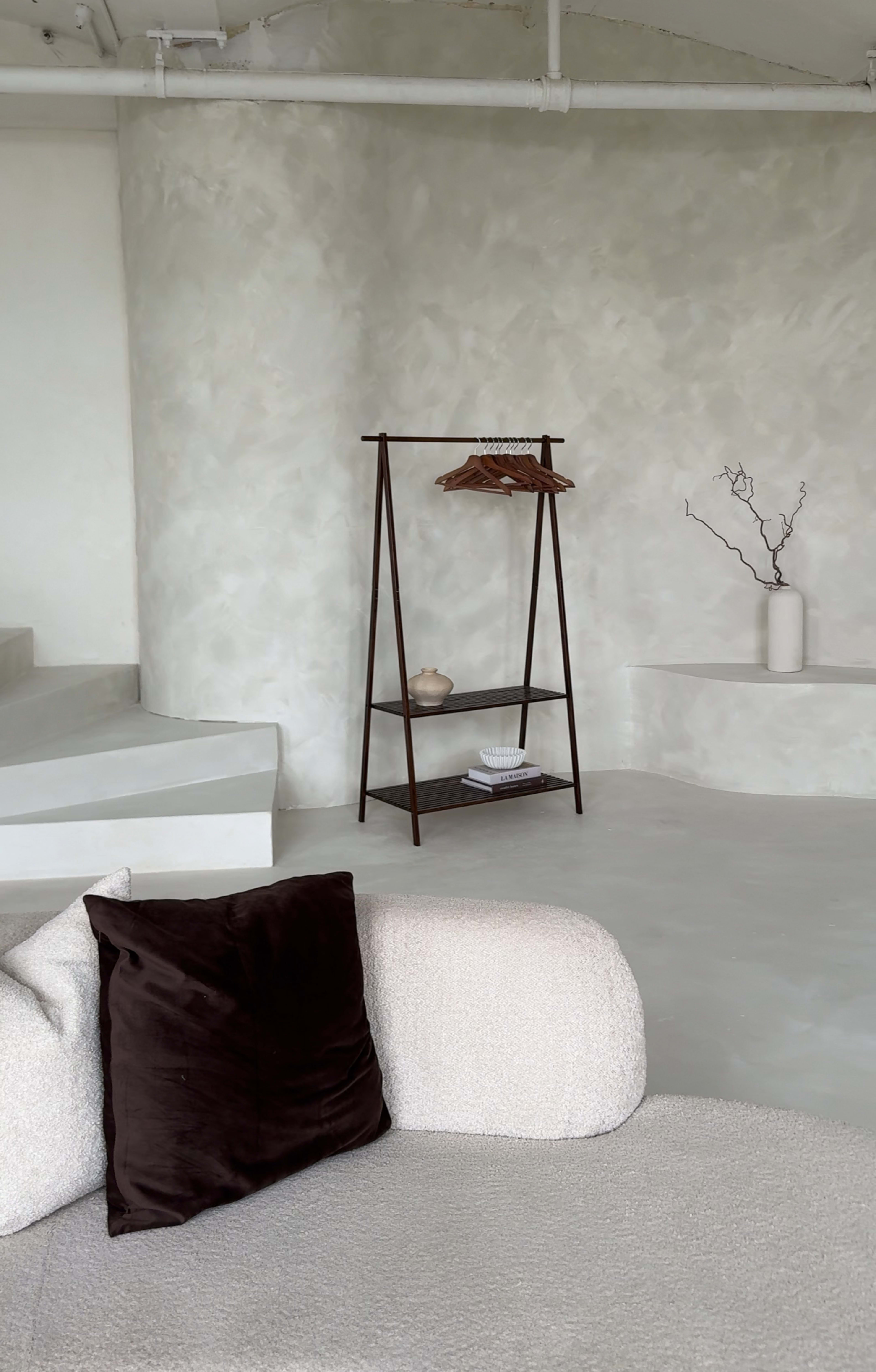 A minimalist interior scene features a light-colored wall, a simple shelving unit with clothing and decorative items, a vase with a branch, and a textured couch in the foreground.