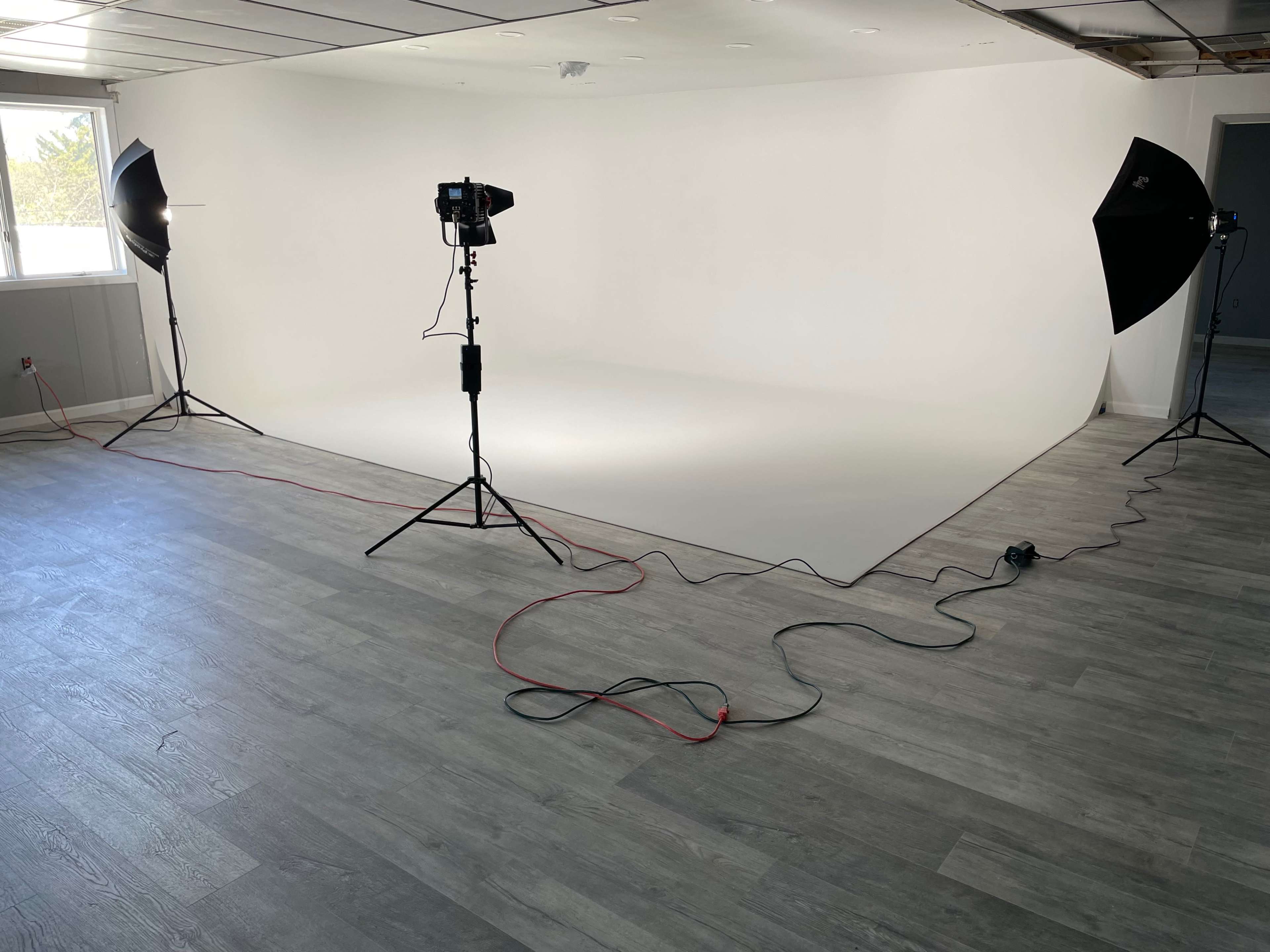 A photography studio with a seamless white backdrop, two light sources on tripods, and cables running across a wooden floor.