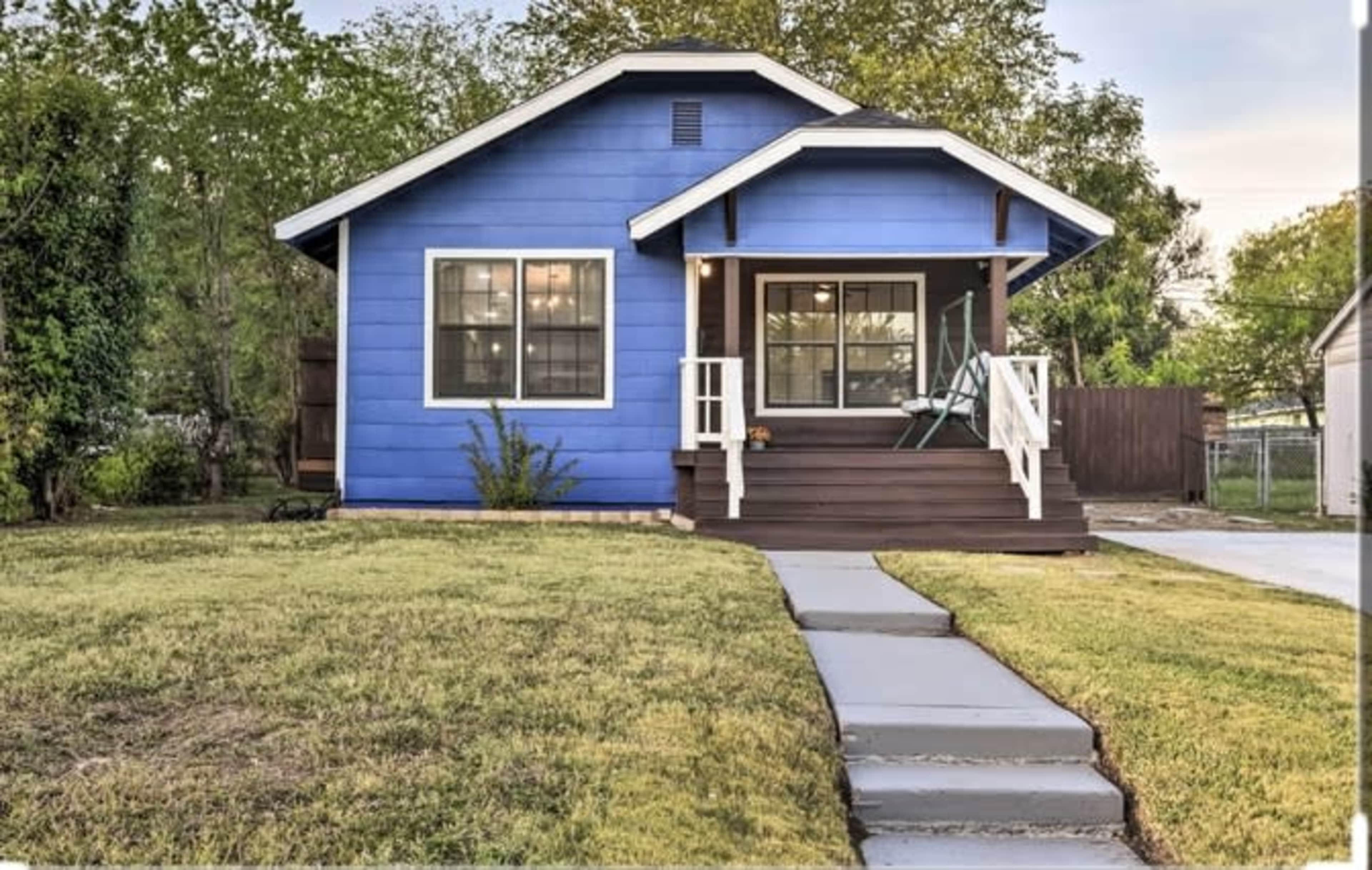 A blue house with white trim features a front porch and a path leading to the entrance, surrounded by grassy areas and trees.