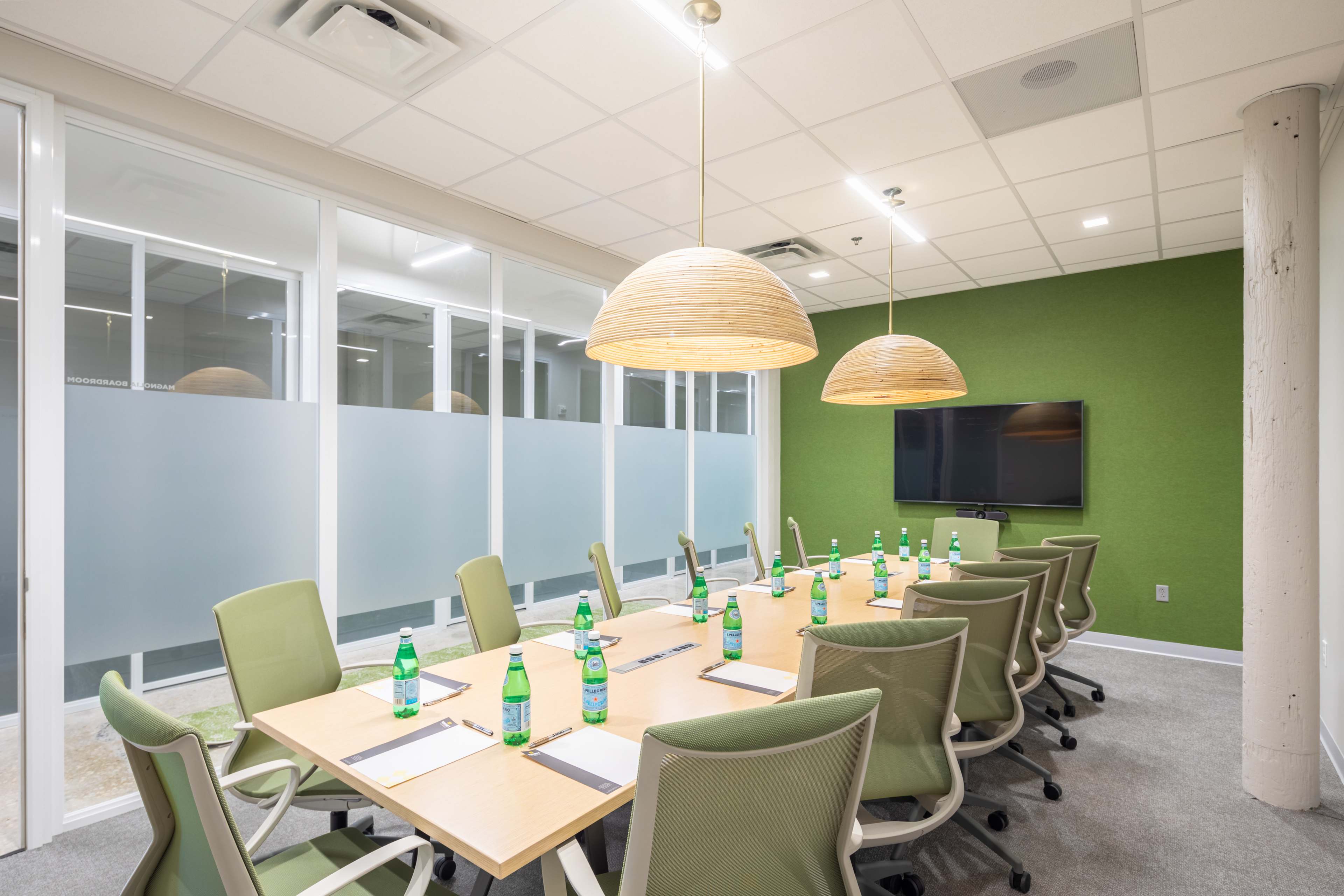 A modern conference room features a long table set with notepads and water bottles, surrounded by green chairs and illuminated by pendant lights.