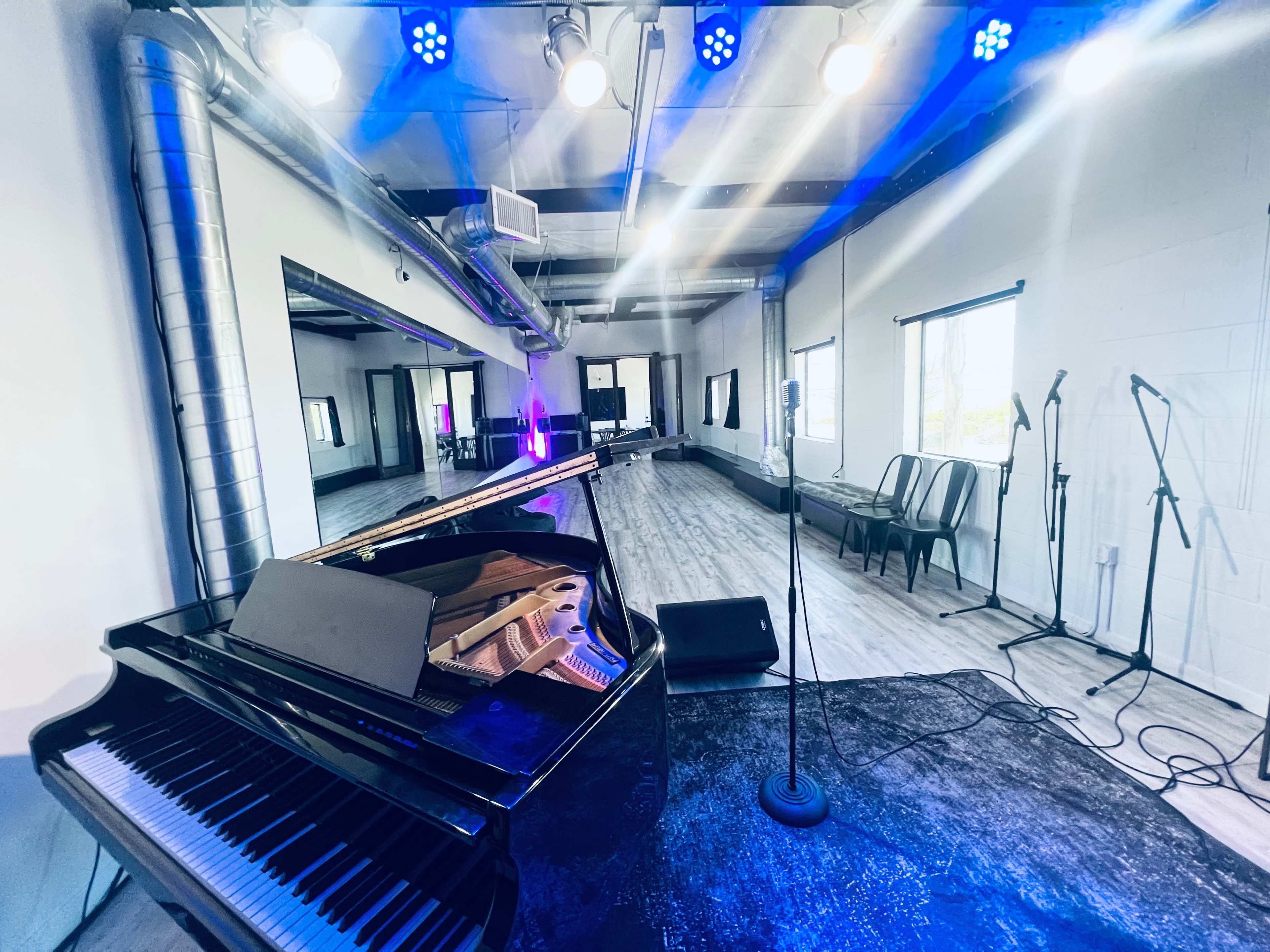 A grand piano sits in the foreground of a well-lit performance space featuring microphones and seating in the background.