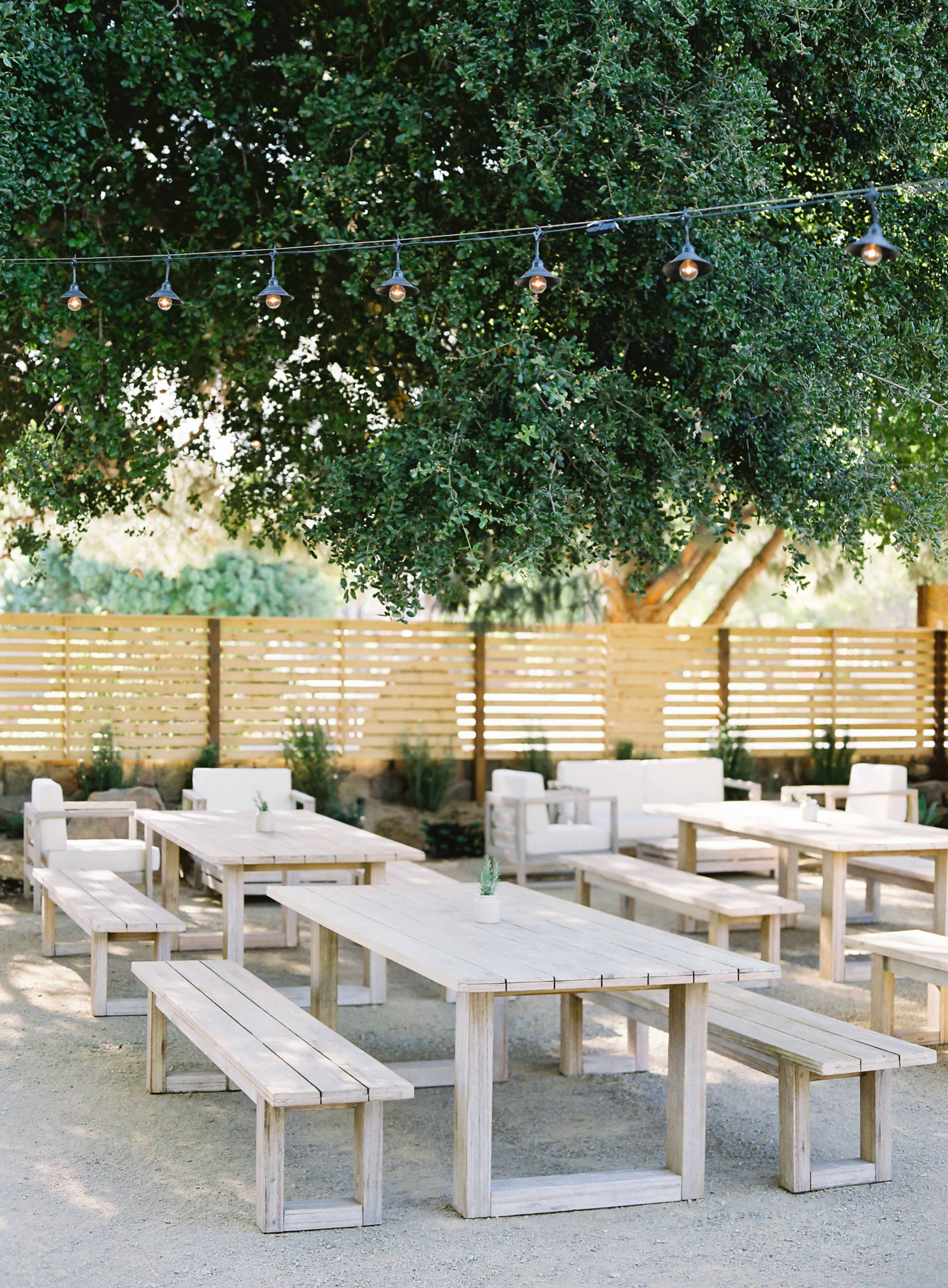 A selection of wooden tables and benches is arranged under a large tree, with string lights overhead and a wooden fence in the background.