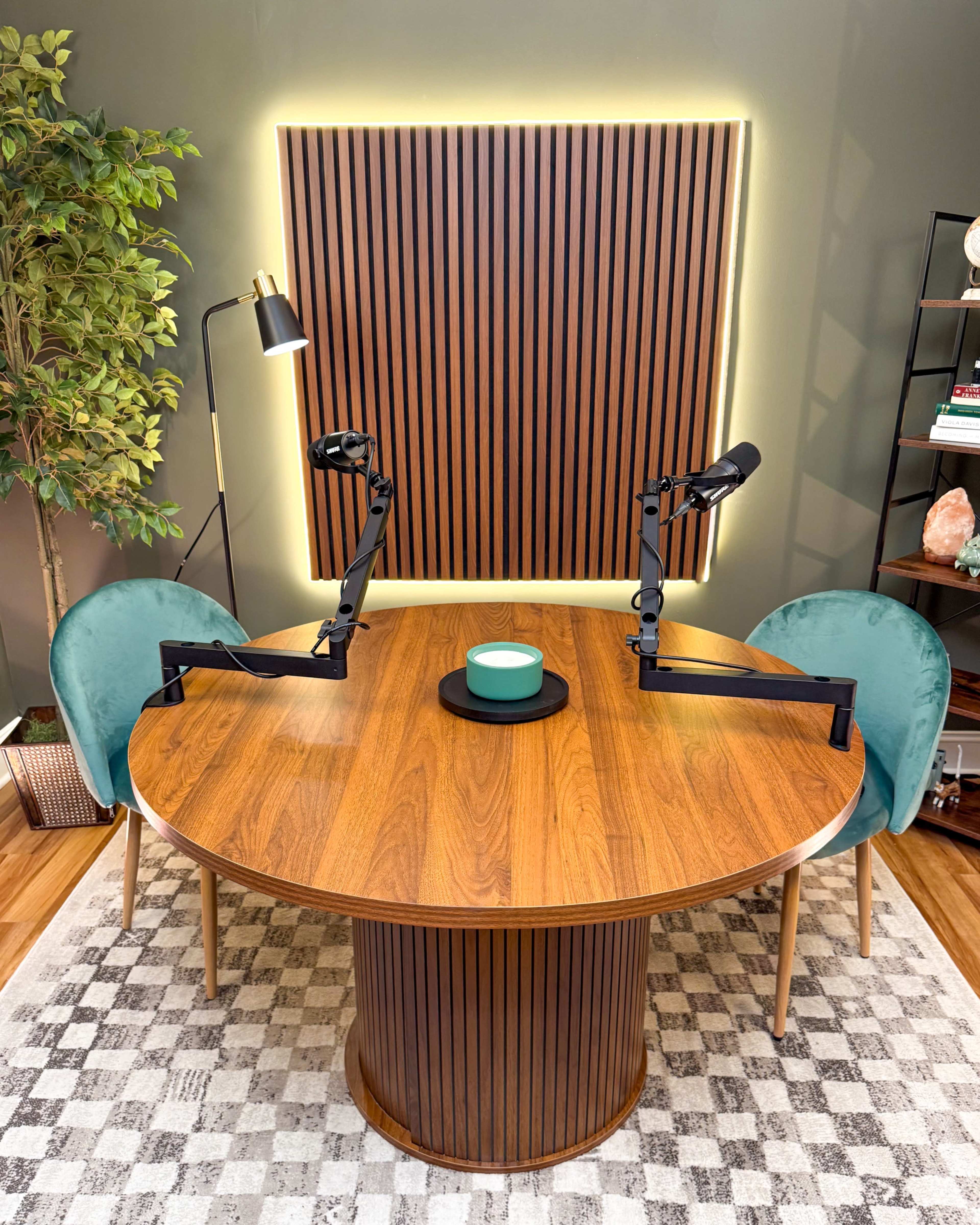 A round wooden table with black microphone arms is set between two teal chairs under a wall with vertical wooden slats illuminated by soft lighting.