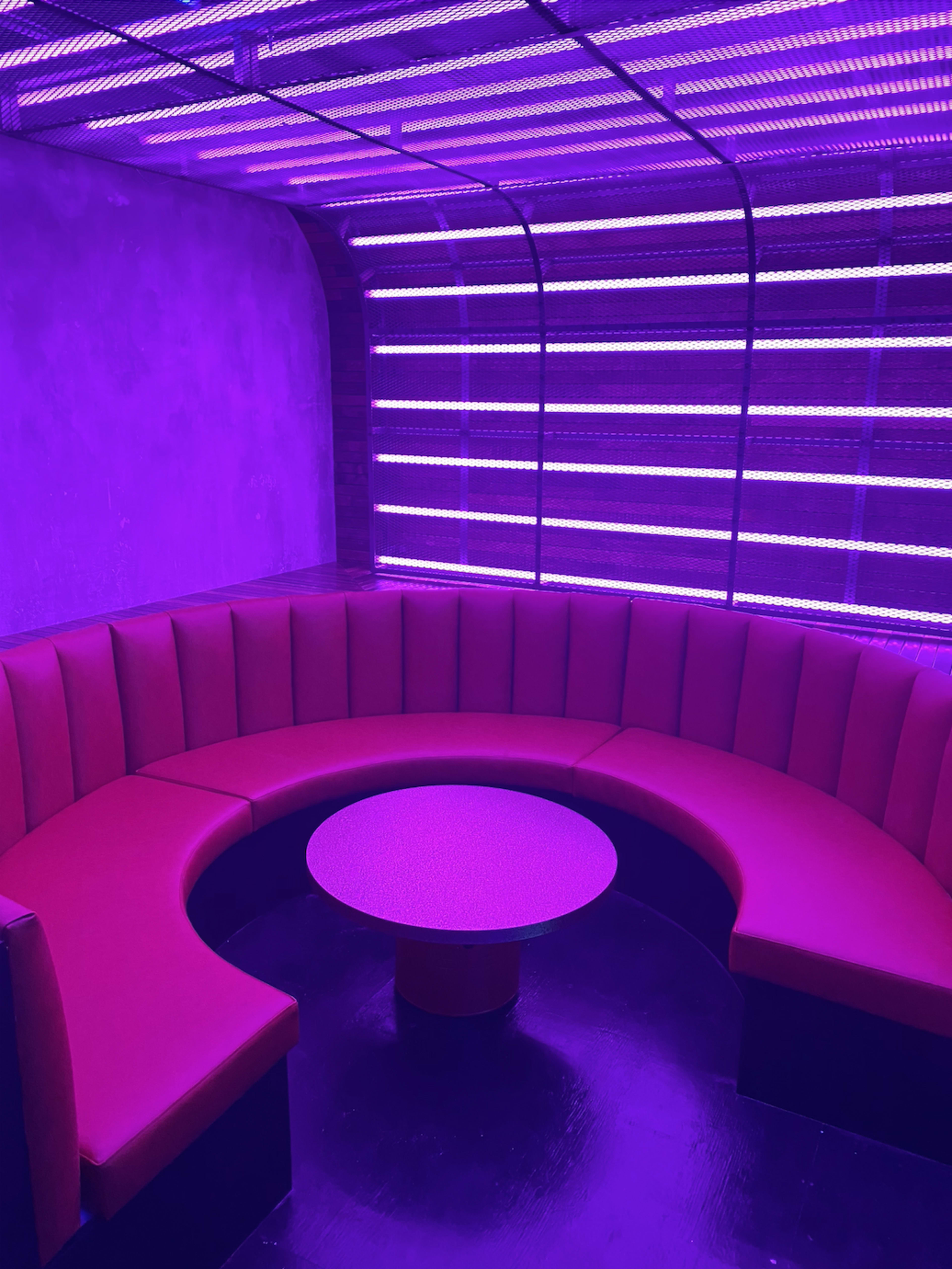 The image shows a dimly lit lounge area featuring a circular red seating arrangement and a round table, all illuminated by purple lighting.