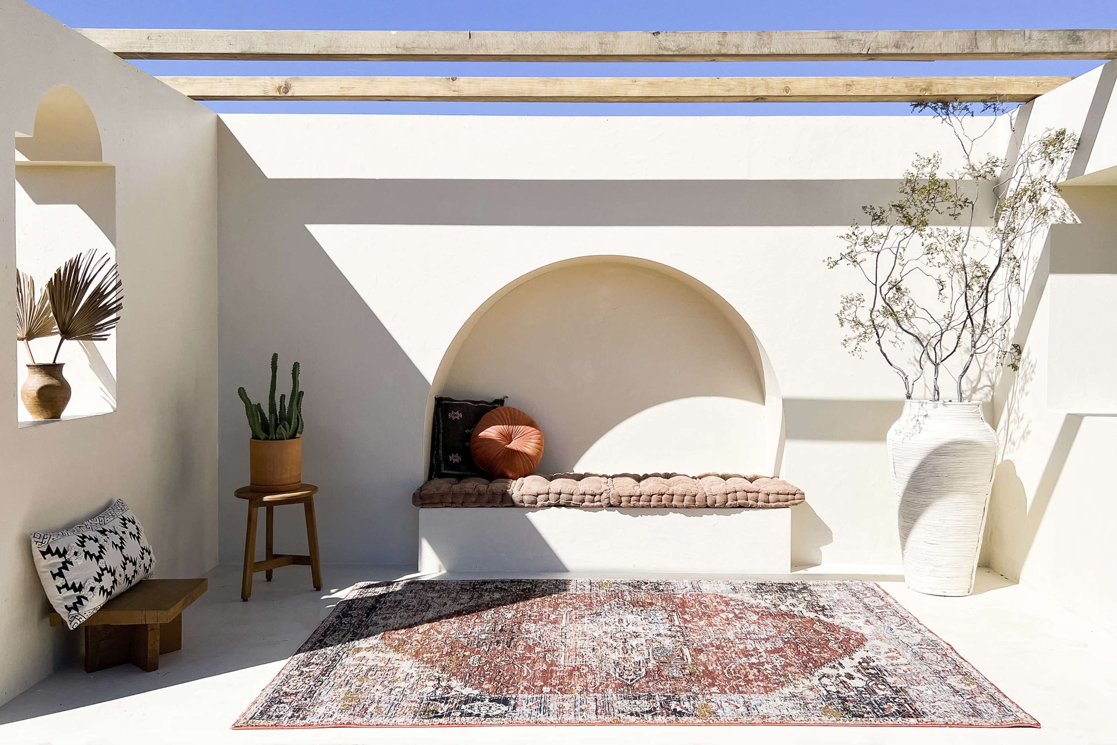 A minimalist indoor space with a curved alcove bench, a decorative rug, a potted plant, and a large vase beside a wall adorned with natural elements.