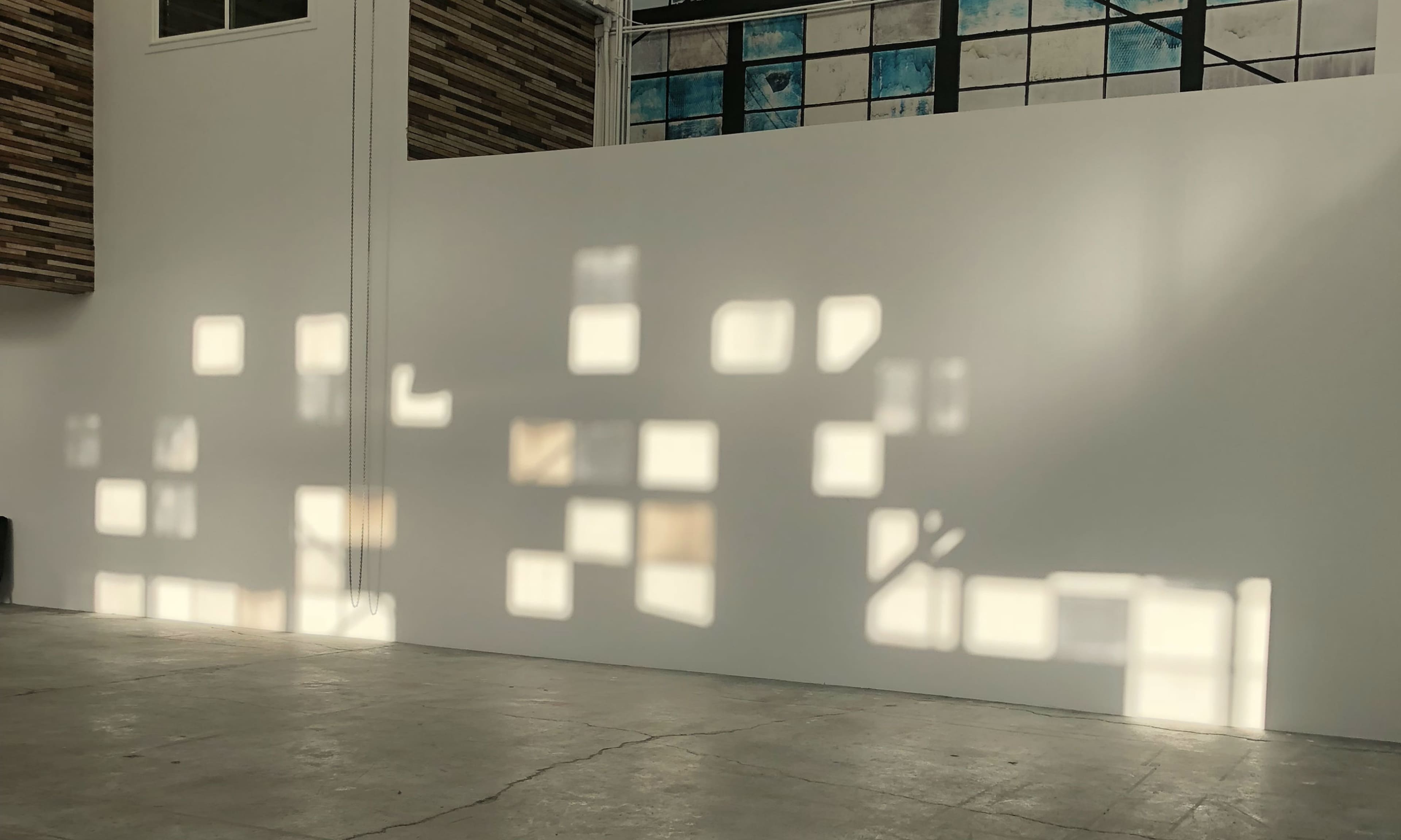 A wall in an industrial space adorned with abstract shadows resembling window panes cast from overhead light.