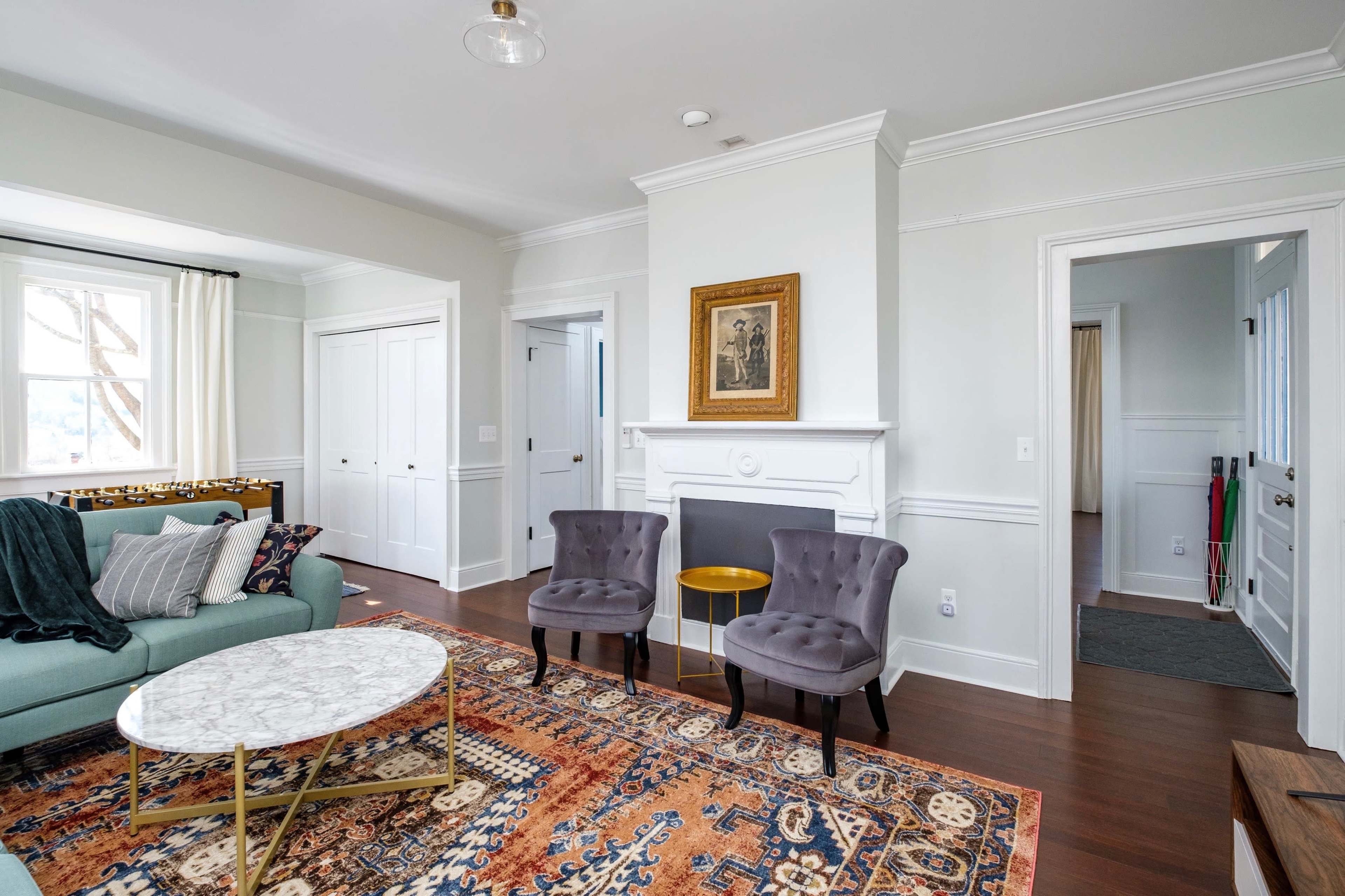 A bright living room with a green couch, two velvet chairs, a marble coffee table, and a decorative area rug, framed by white wainscoting and doorways leading to other rooms.