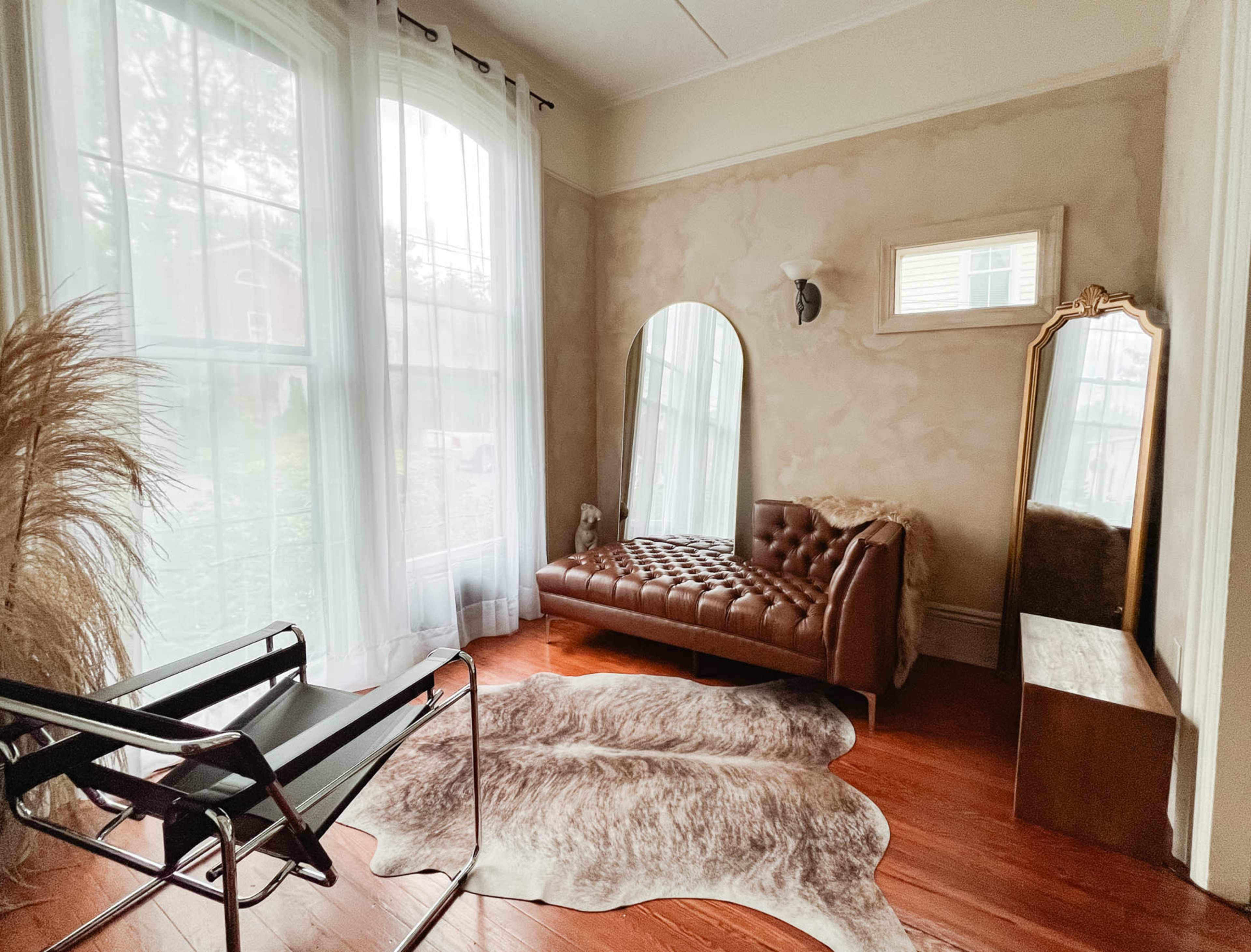 A corner of a room featuring a brown leather tufted sofa, a large mirror, a chair, and a cowhide rug on wooden flooring beside tall windows with sheer curtains.