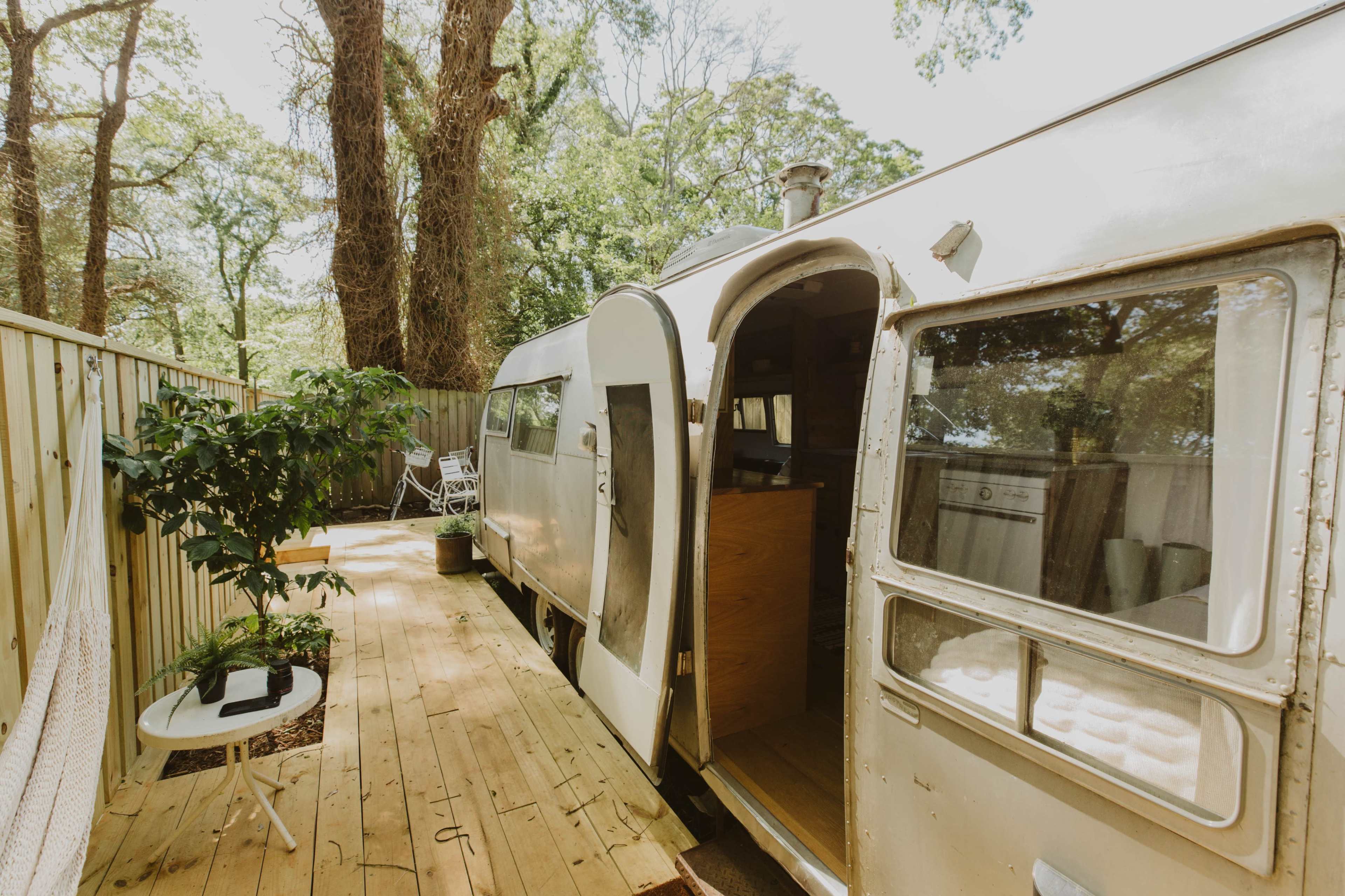 An airstream trailer is parked on a wooden deck surrounded by trees, with a door open revealing the interior.