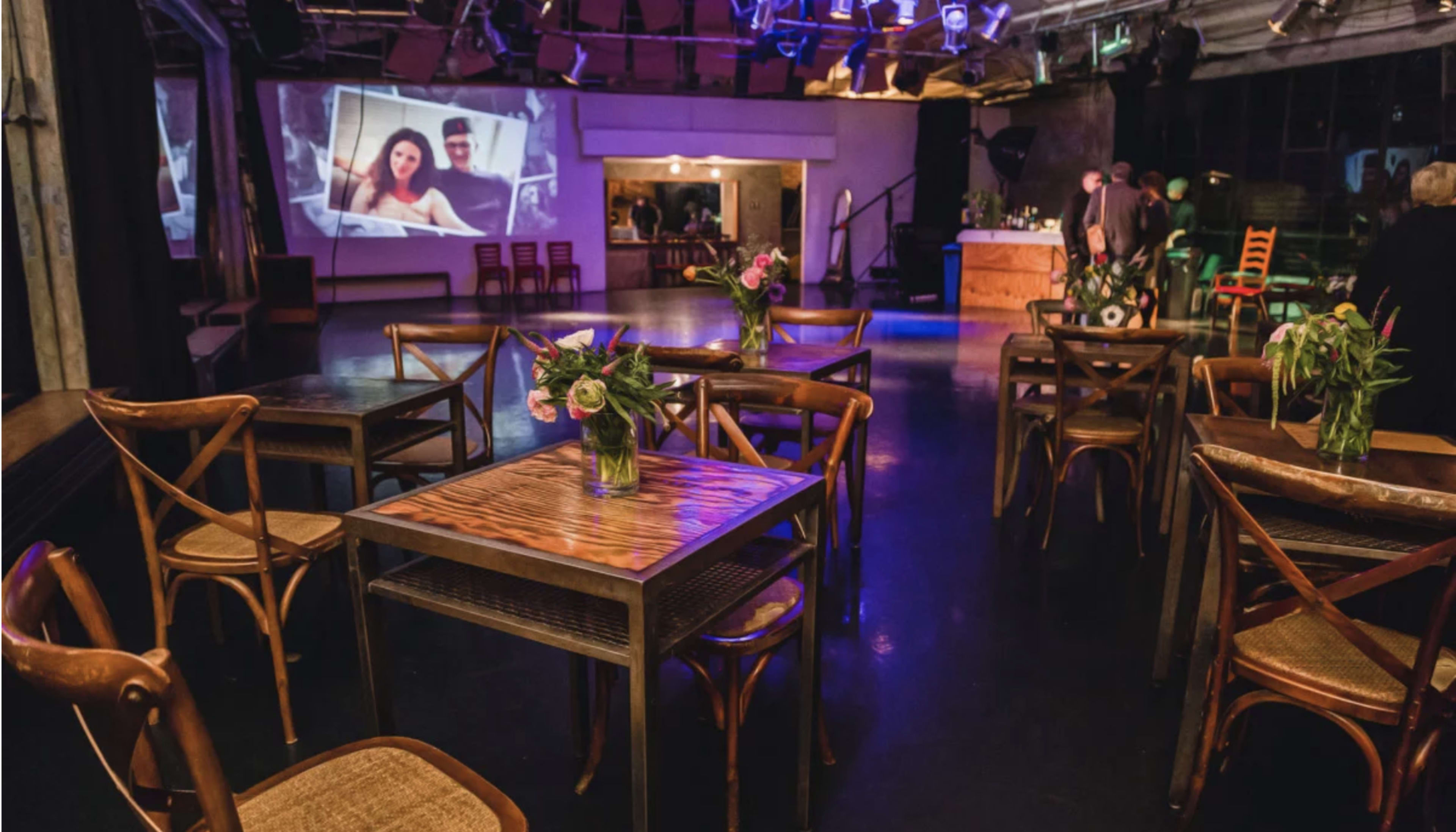 The image shows a modern event space with wooden tables and chairs arranged around a room featuring a projection screen displaying a couple.