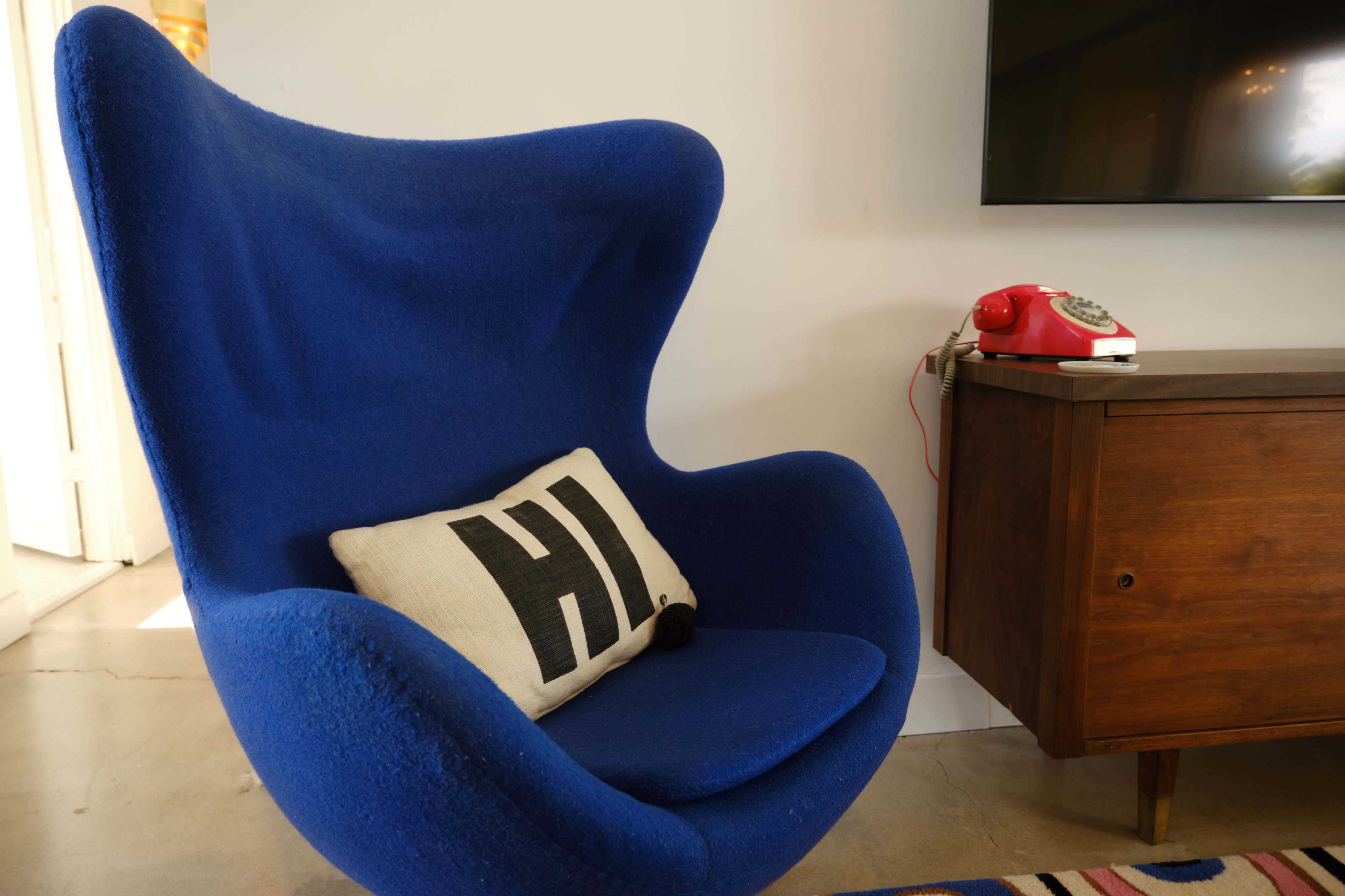 A blue, modern armchair next to a wooden cabinet and a vintage red telephone, with a cushion that has "HI" printed on it.