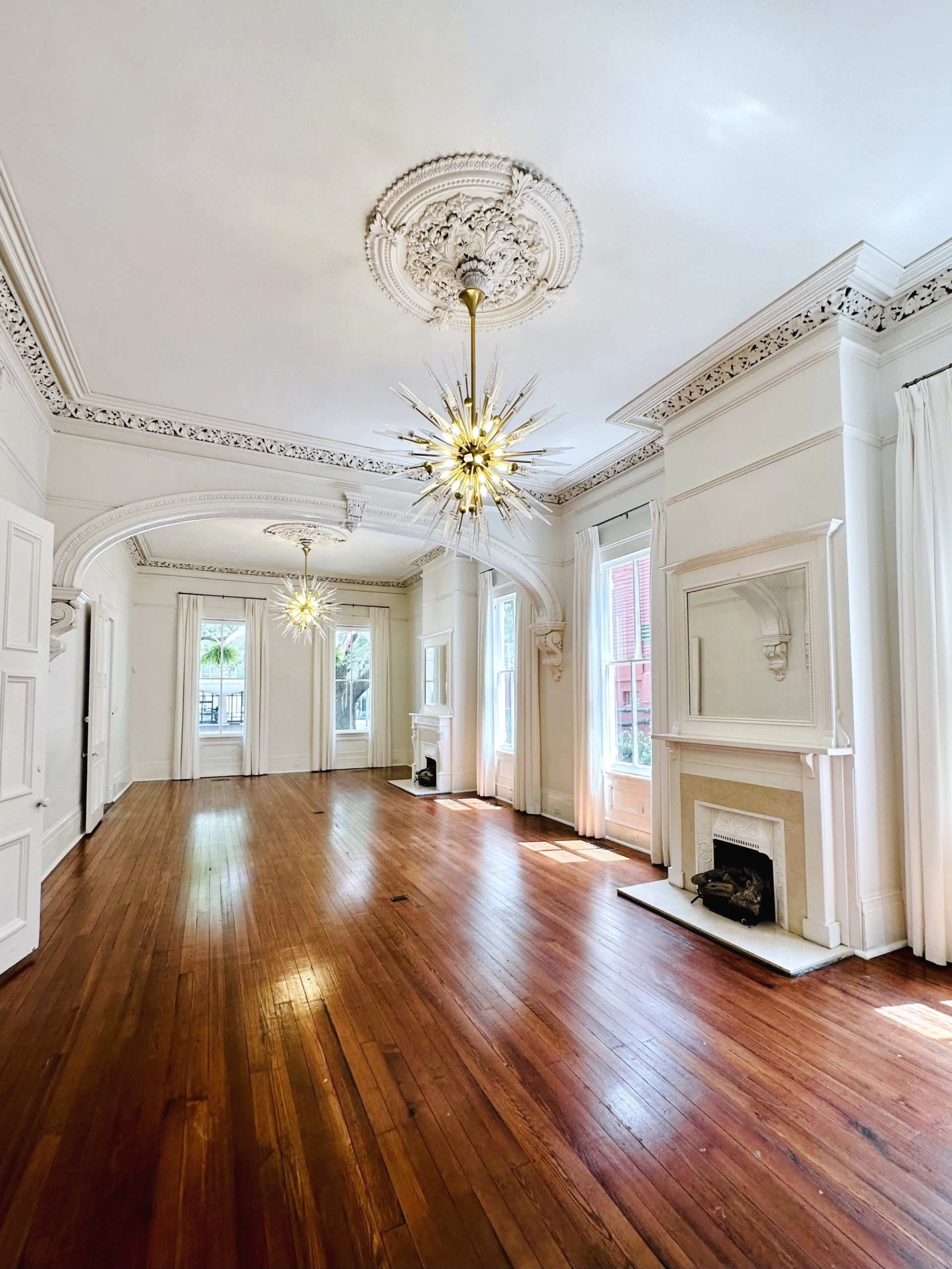 A spacious room features hardwood floors, intricate ceiling moldings, and two ornate chandeliers.