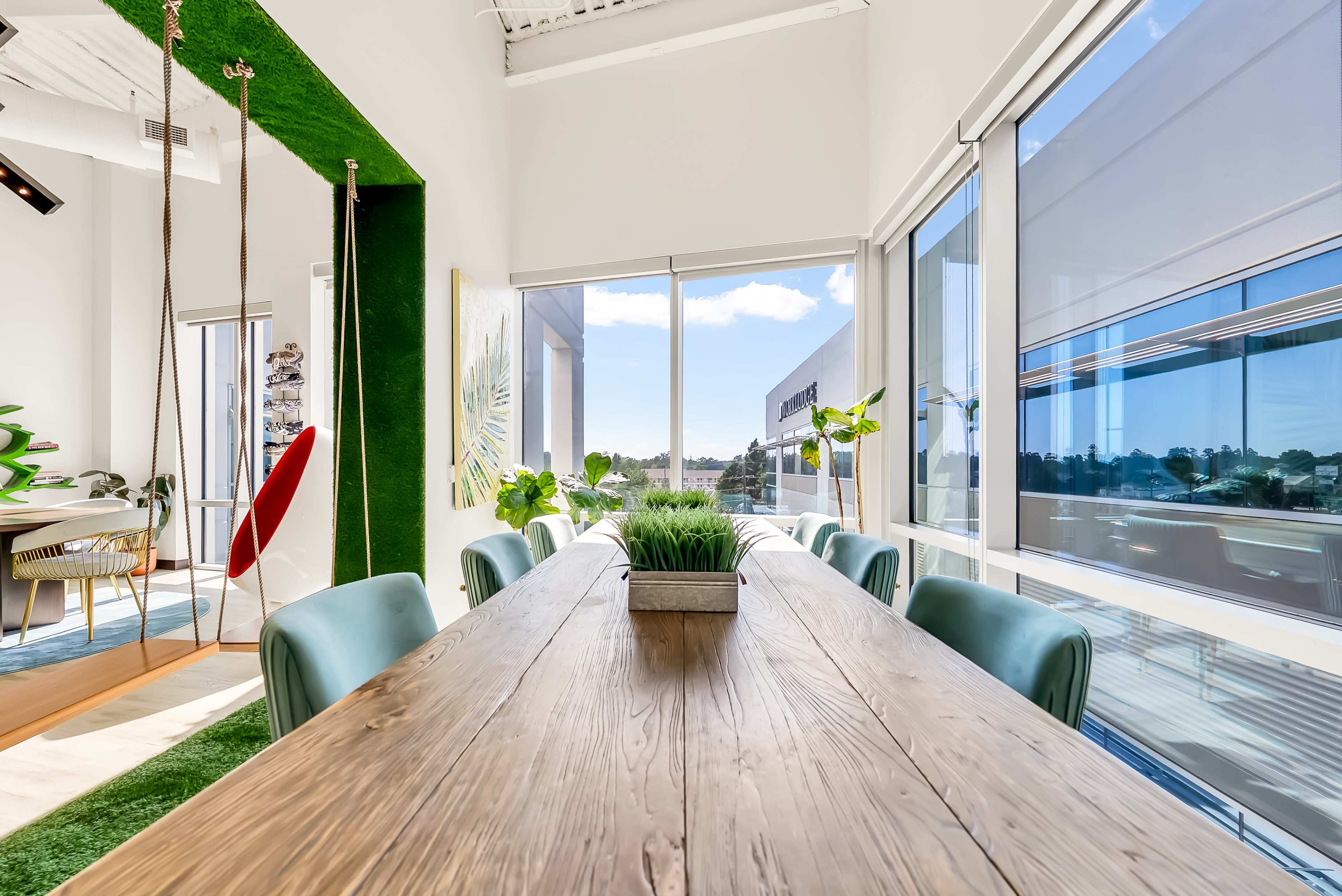 A long wooden dining table is set in a bright room with large windows, showcasing greenery outside and decorated with modern furnishings.