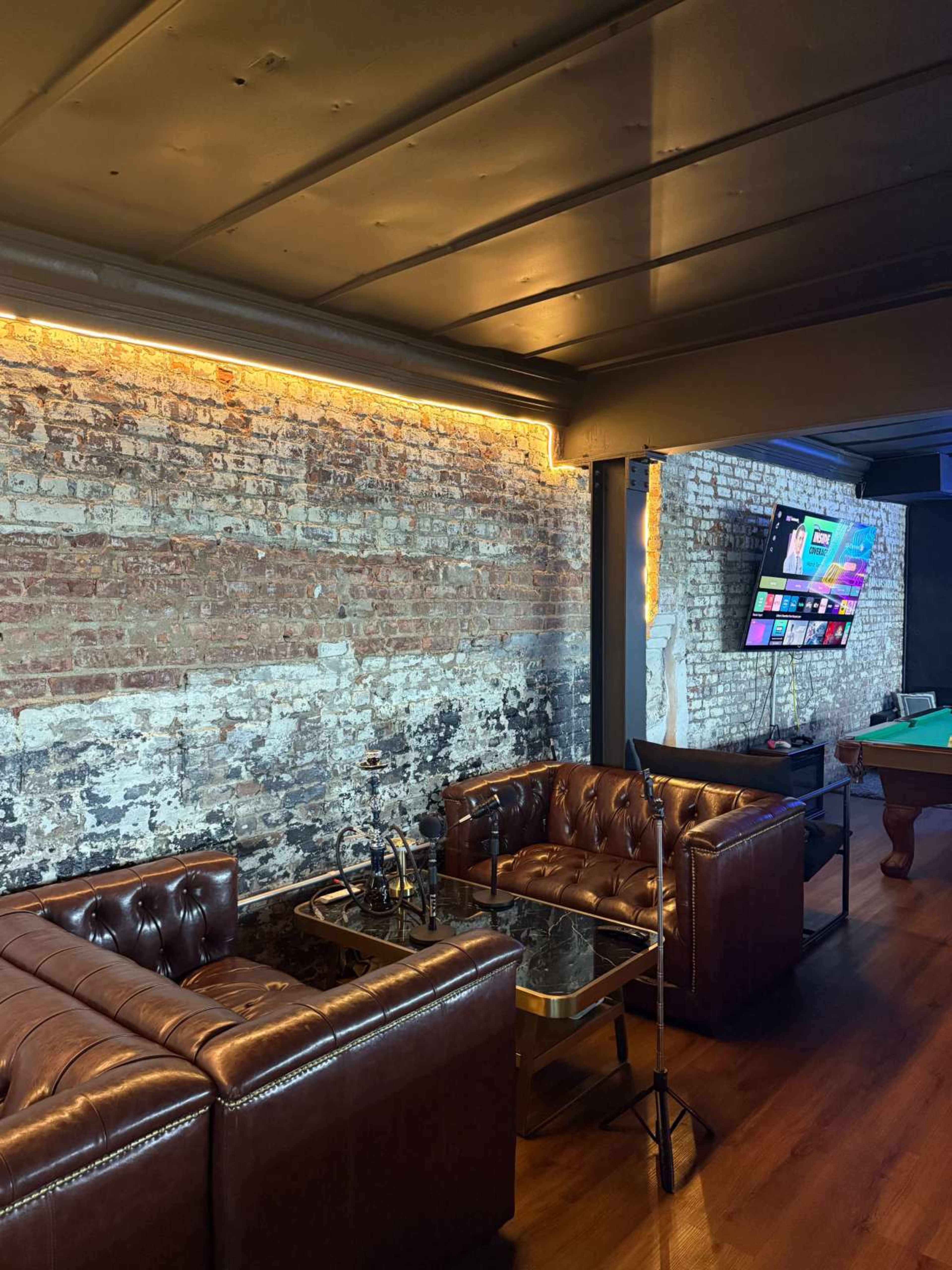 The image shows a cozy lounge area with brown leather sofas, a glass coffee table, a pool table, and a wall featuring exposed brick and modern lighting.