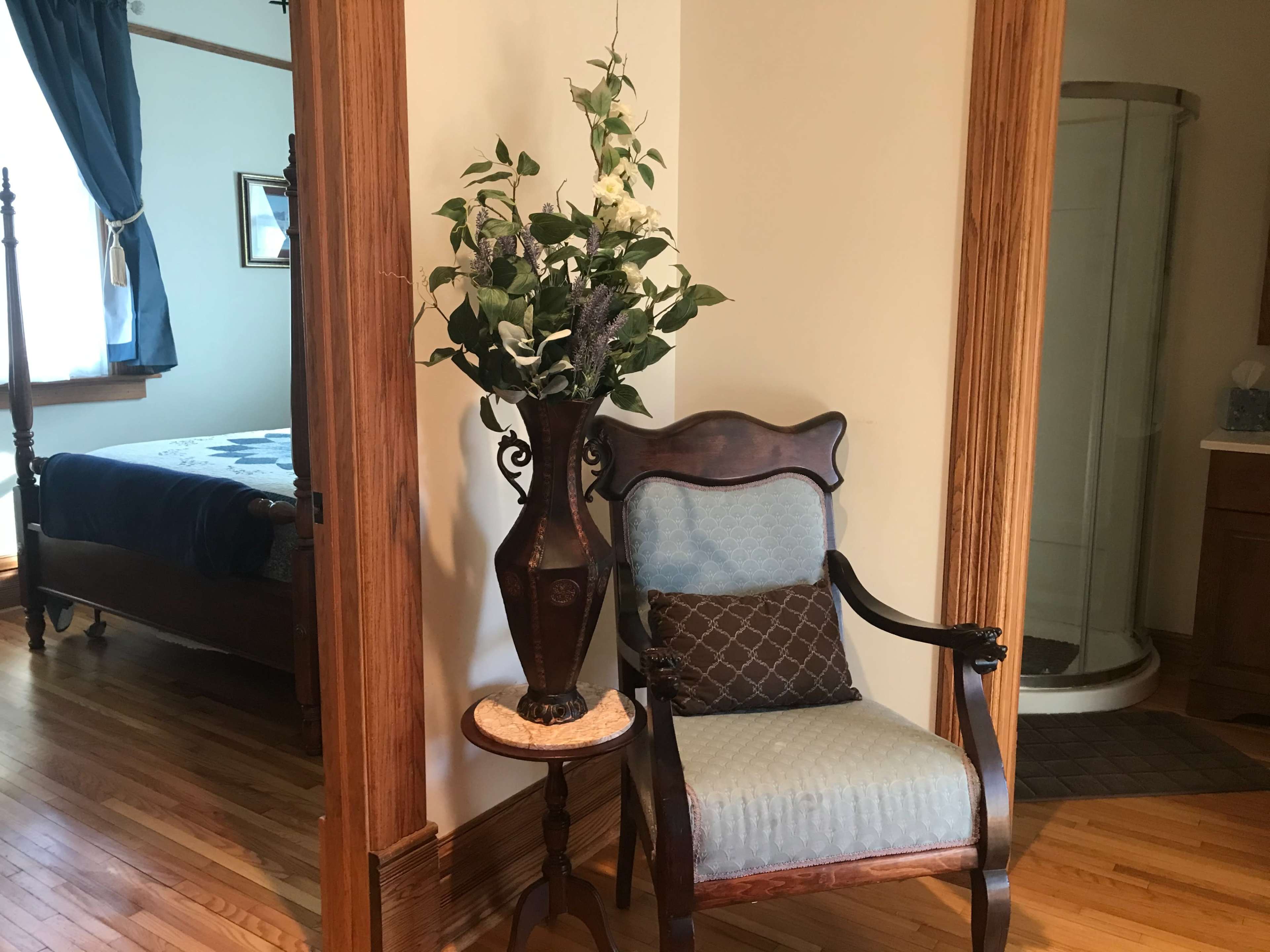 A wooden chair beside a decorative plant in a vase, framed by a doorway leading to a bedroom and a bathroom area.