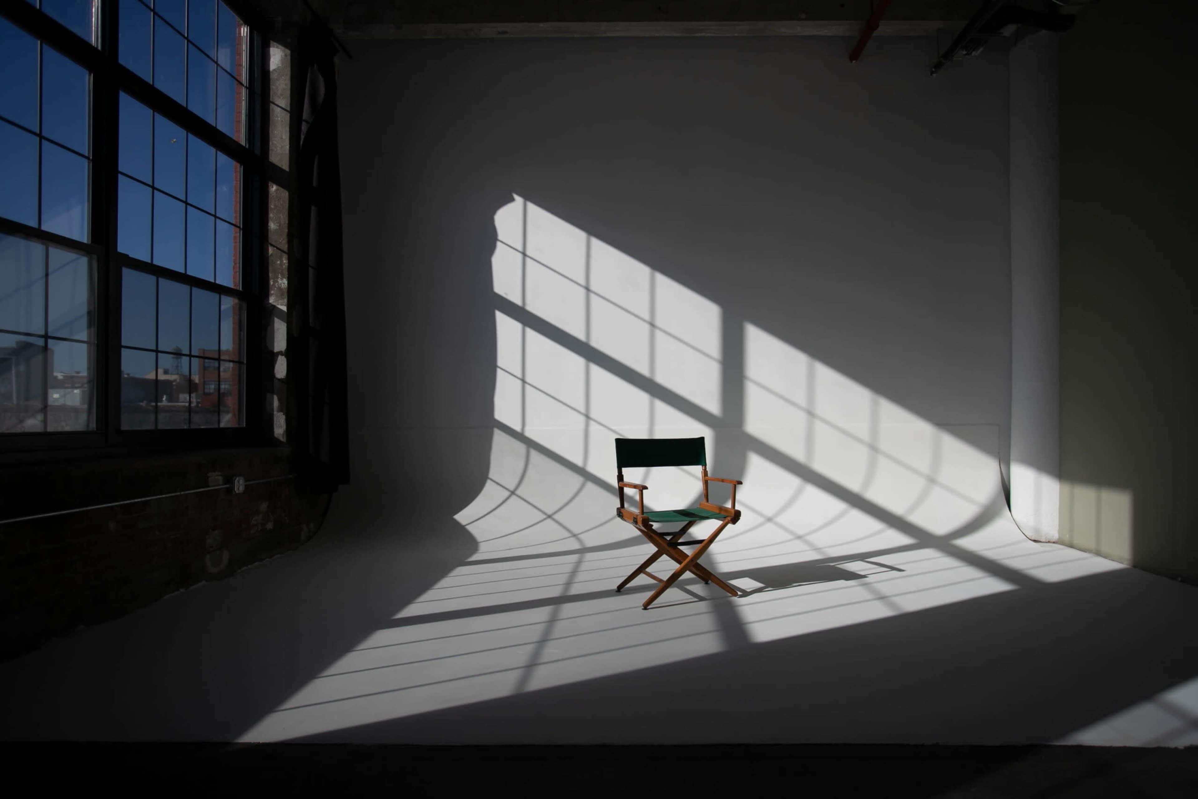 A wooden director's chair sits on a white studio floor, illuminated by sunlight casting geometric shadows from large windows.