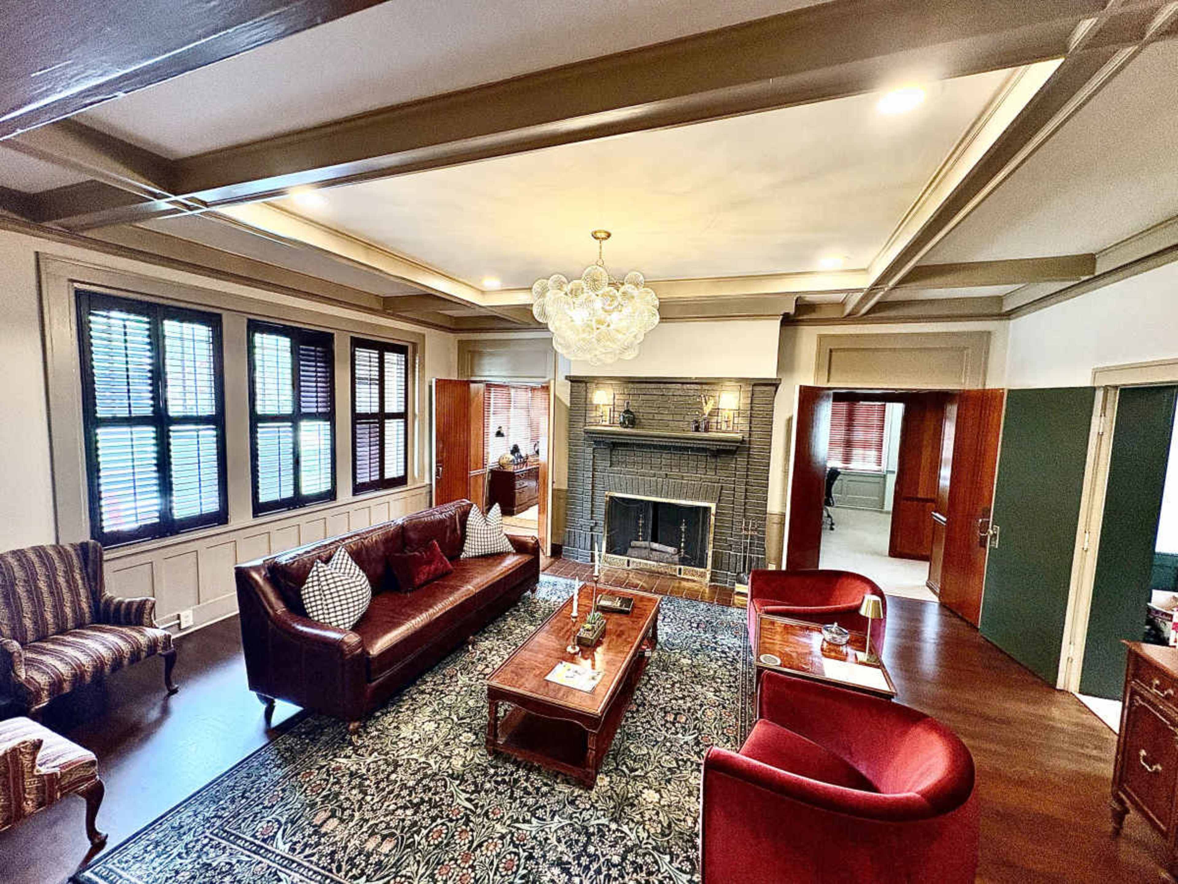 The image shows a well-furnished living room with a central coffee table, two red chairs, a brown sofa, a fireplace, and large windows with shutters.