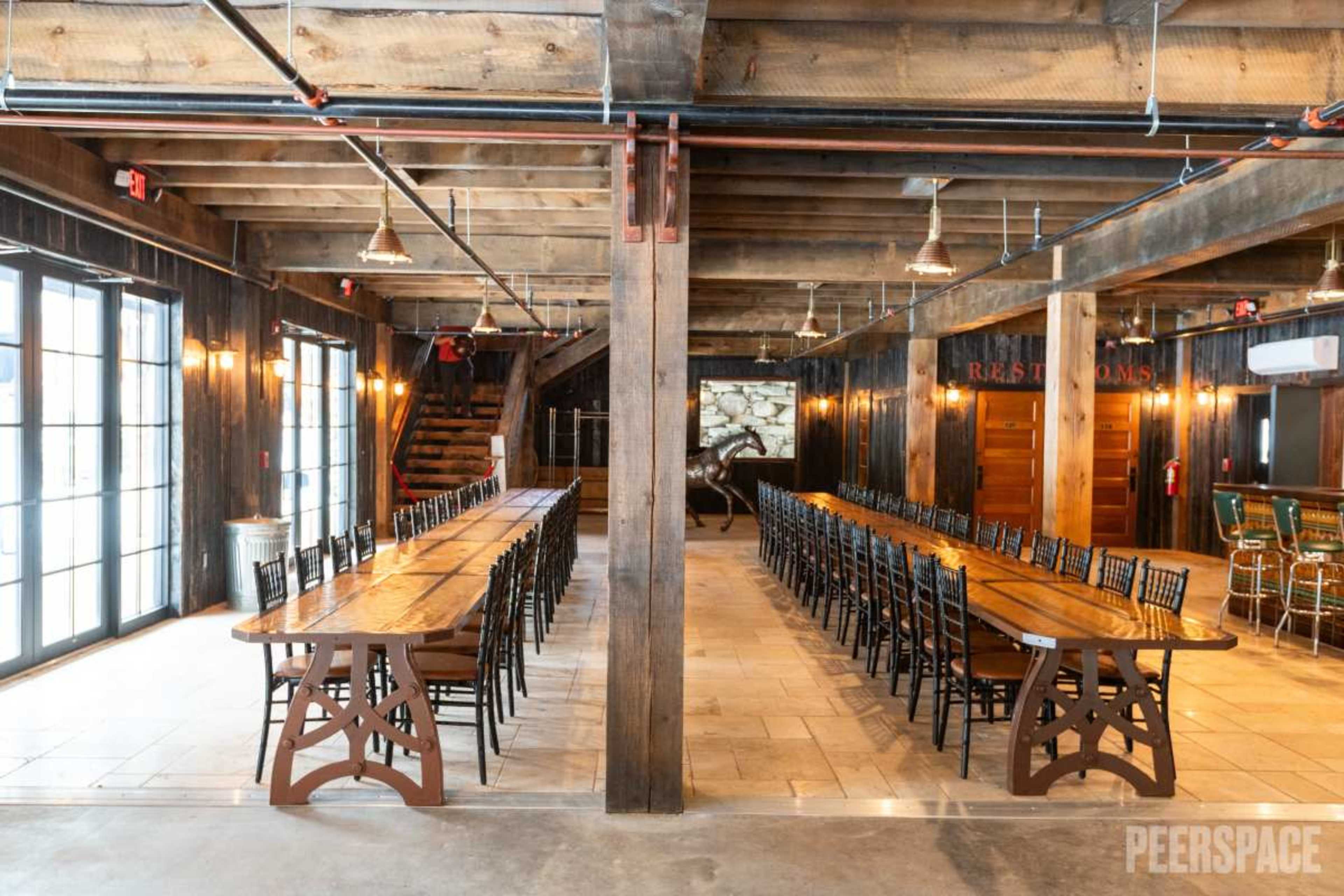 The image shows a spacious dining area with large wooden tables arranged in a U-shape, surrounded by high-backed chairs, and features exposed wooden beams and industrial lighting.