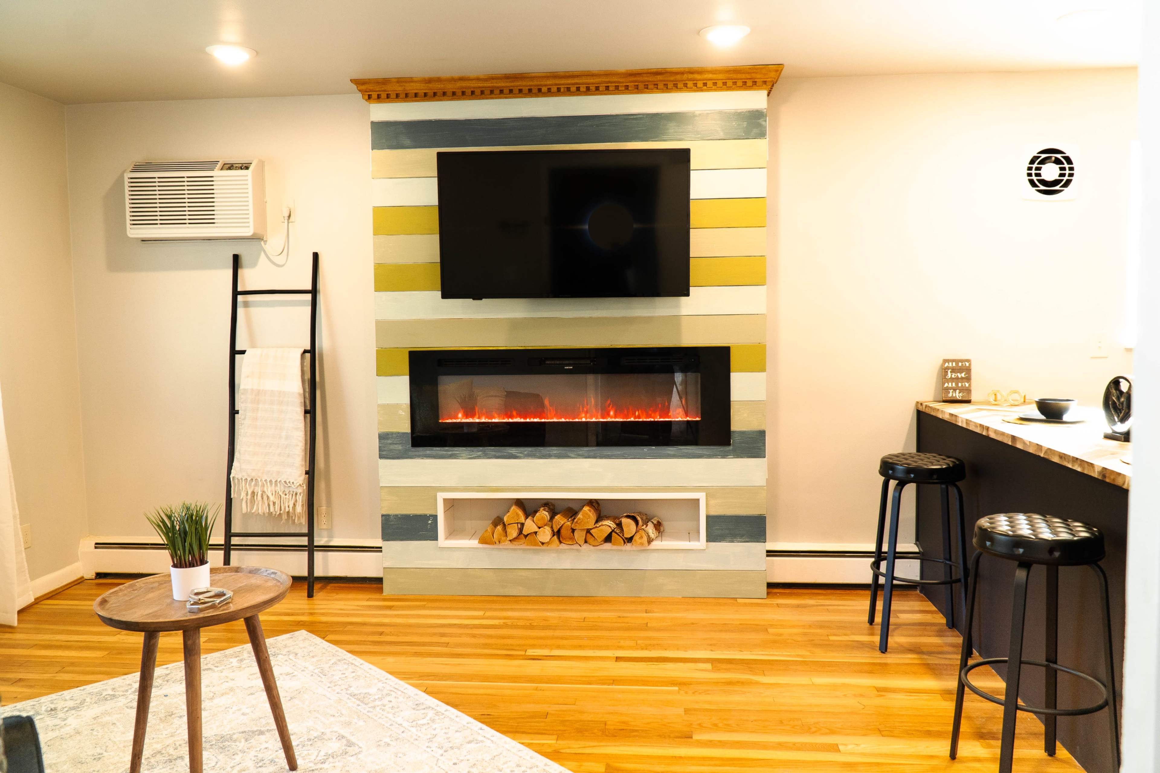 A modern living area with a striped wall featuring a mounted TV and an electric fireplace, complemented by a wooden table and bar stools.